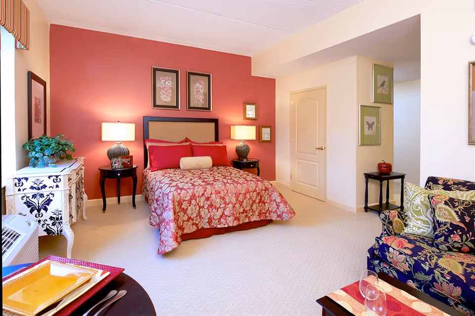 A cozy bedroom with a bed featuring a floral red and pink bedspread and matching pillows against a red accent wall. Two bedside tables with lamps flank the bed. The room also includes a white decorative dresser with a green plant, framed artwork on the walls, a floral patterned armchair with green and floral cushions, and a small table with a red decorative item. The carpet is light beige, and there is a window with a striped valance.