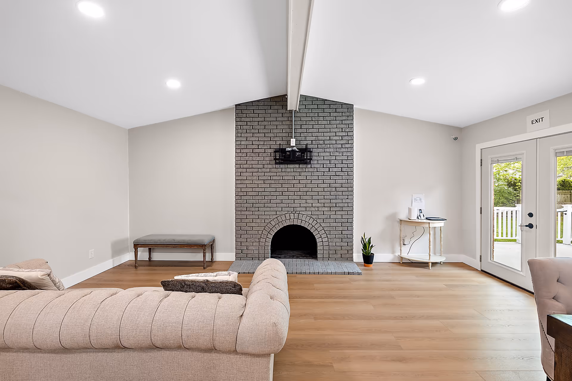 A bright living room with light wood flooring, a beige tufted sofa, a gray brick fireplace centered on the far wall, a small bench to the left, a small table with a plant and papers to the right, and double glass doors leading outside with an exit sign above.