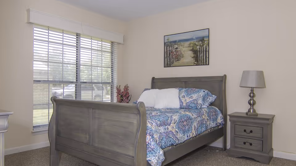 A bright bedroom featuring a wooden sleigh bed with patterned bedding, a nightstand and lamp, a large window, and wall art.
