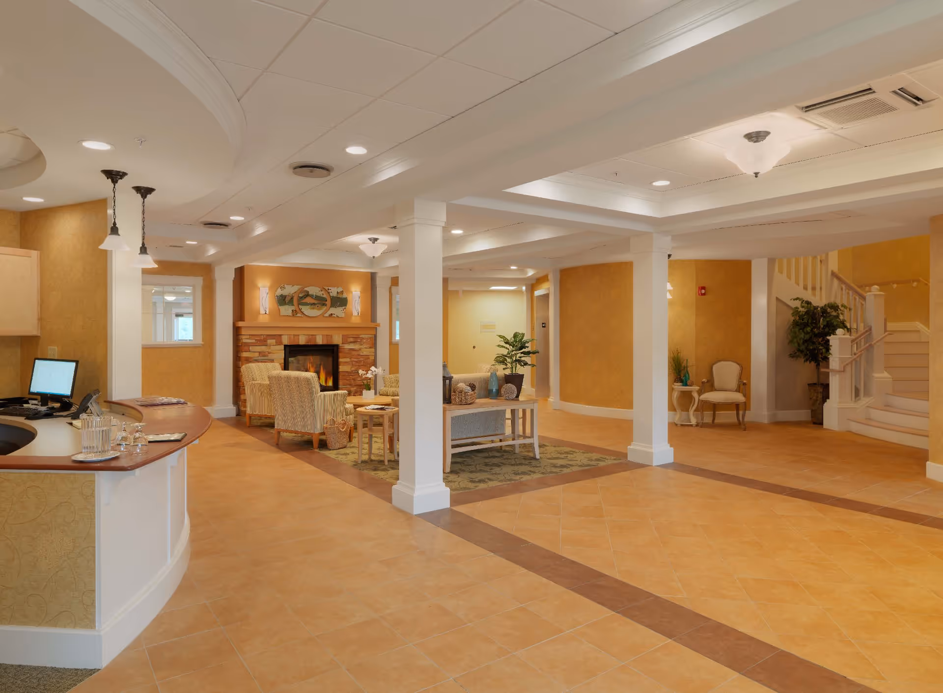 Warm, spacious senior living facility lobby with a reception desk, seating area by a fireplace, and a staircase.