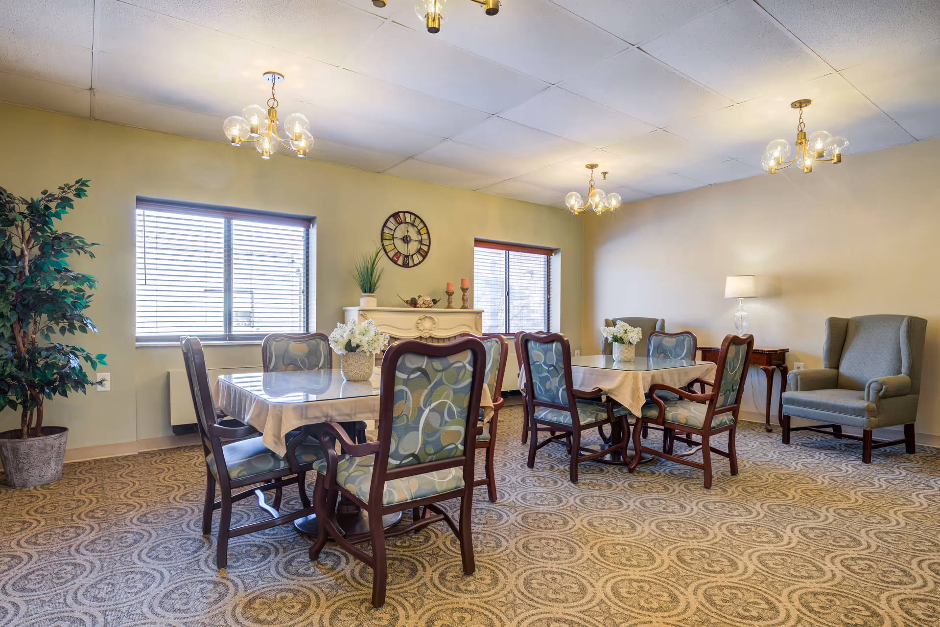 A cozy dining room with two tables covered with tablecloths, each surrounded by four wooden chairs with patterned cushions. There are two large windows with blinds, a decorative clock on the wall, a potted plant, and a white mantelpiece with decorative items. The room has patterned carpet flooring, two armchairs, a side table with a lamp, and three ceiling light fixtures.