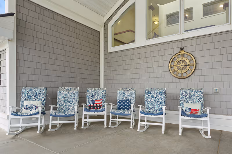 Six white rocking chairs with blue patterned cushions and patriotic-themed pillows are arranged in a row on a covered porch with gray shingle siding and a large decorative clock on the wall.