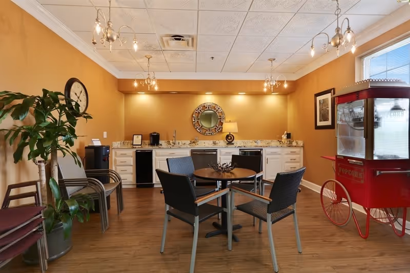 Warm, well-lit communal lounge with round table, chairs, kitchenette, and popcorn cart.