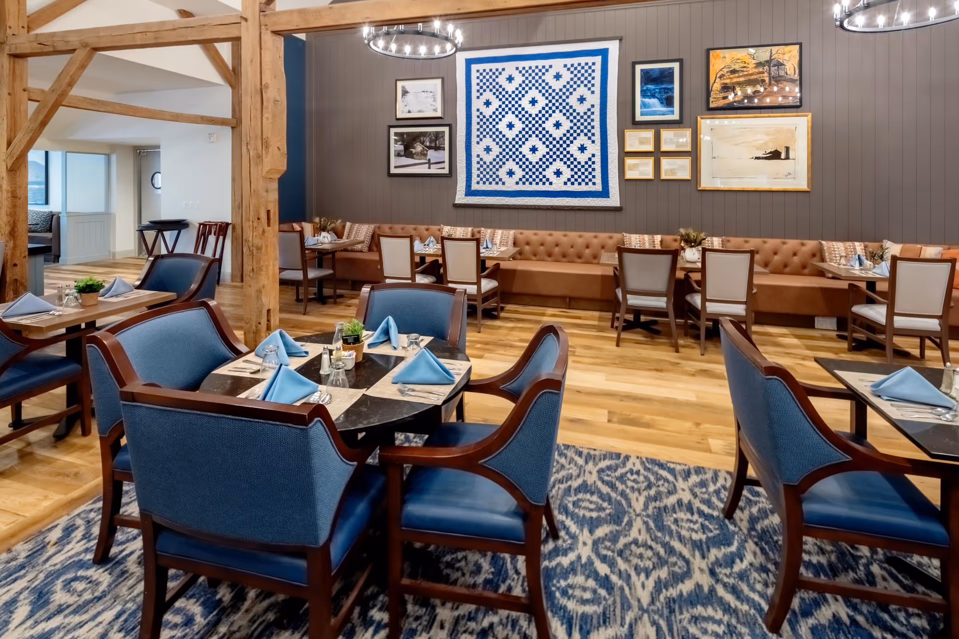 A dining room in a senior living facility with wooden floors, blue upholstered chairs around tables set with blue napkins, and a long tufted bench along the wall. The wall is decorated with framed artwork and a large blue and white quilt. Wooden beams and modern circular chandeliers are visible in the ceiling.