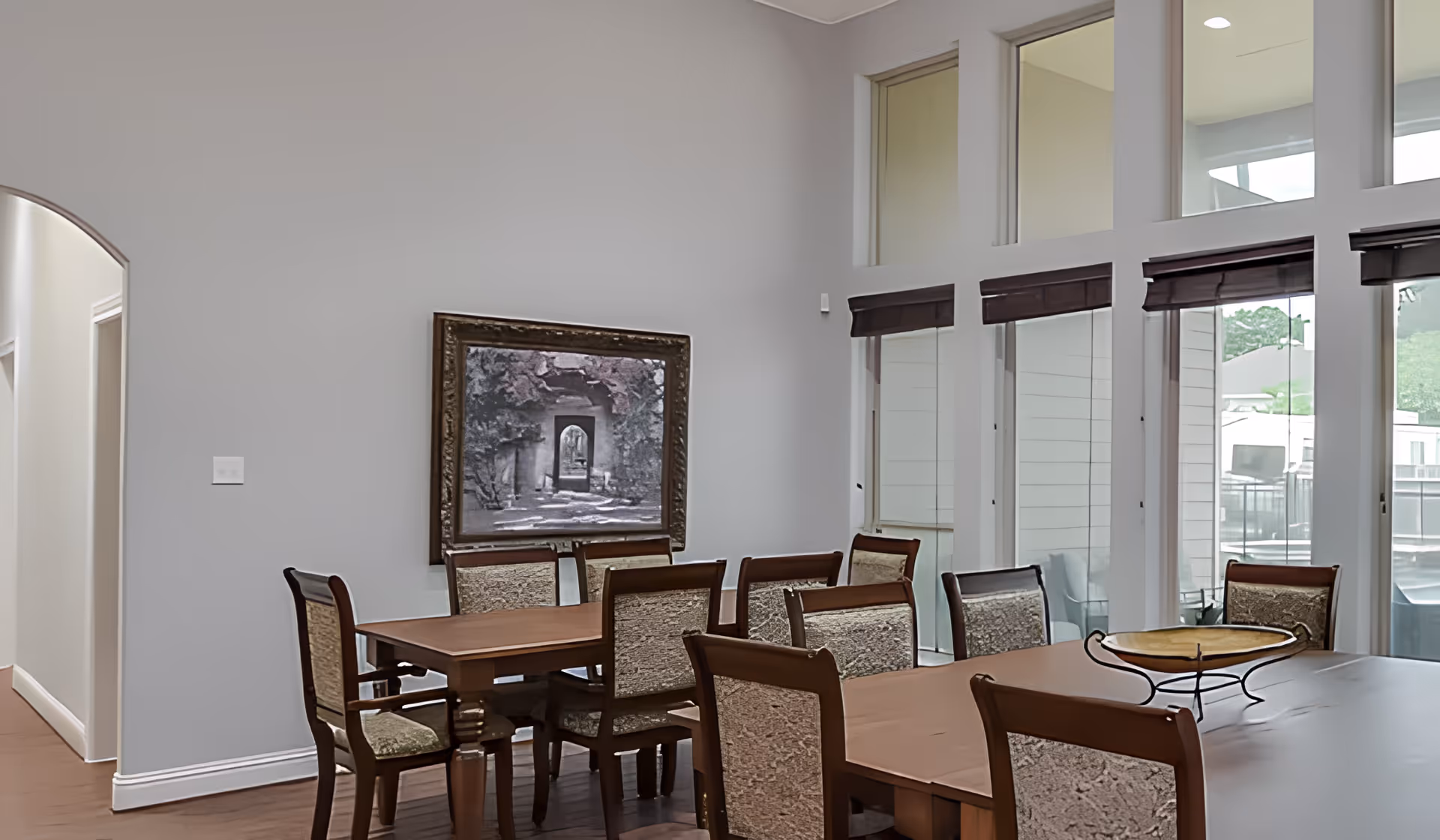 A dining room with two wooden tables and several cushioned chairs. The room has high ceilings with large windows covered by dark blinds, allowing natural light to enter. A framed picture hangs on the light gray wall, and a decorative bowl sits on one of the tables. The floor is wooden, and there is an arched doorway leading to another area.