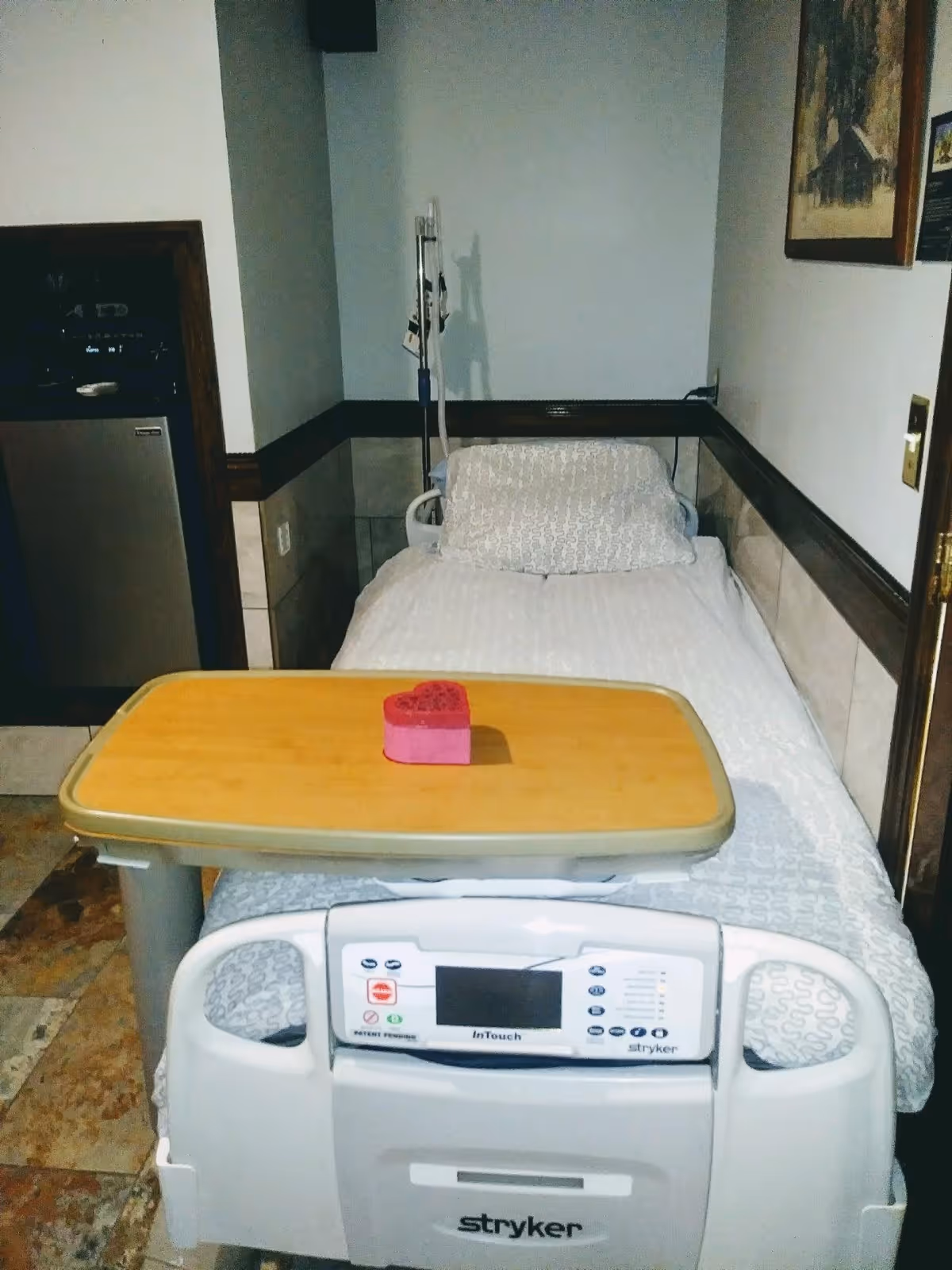 A hospital-style bed with white patterned bedding and a pillow, positioned against a wall in a small room. There is an adjustable overbed table with a pink heart-shaped object on it. An IV pole with bags is visible behind the bed. The room has tiled flooring and a small refrigerator or cabinet in the corner. A framed picture hangs on the wall to the right.