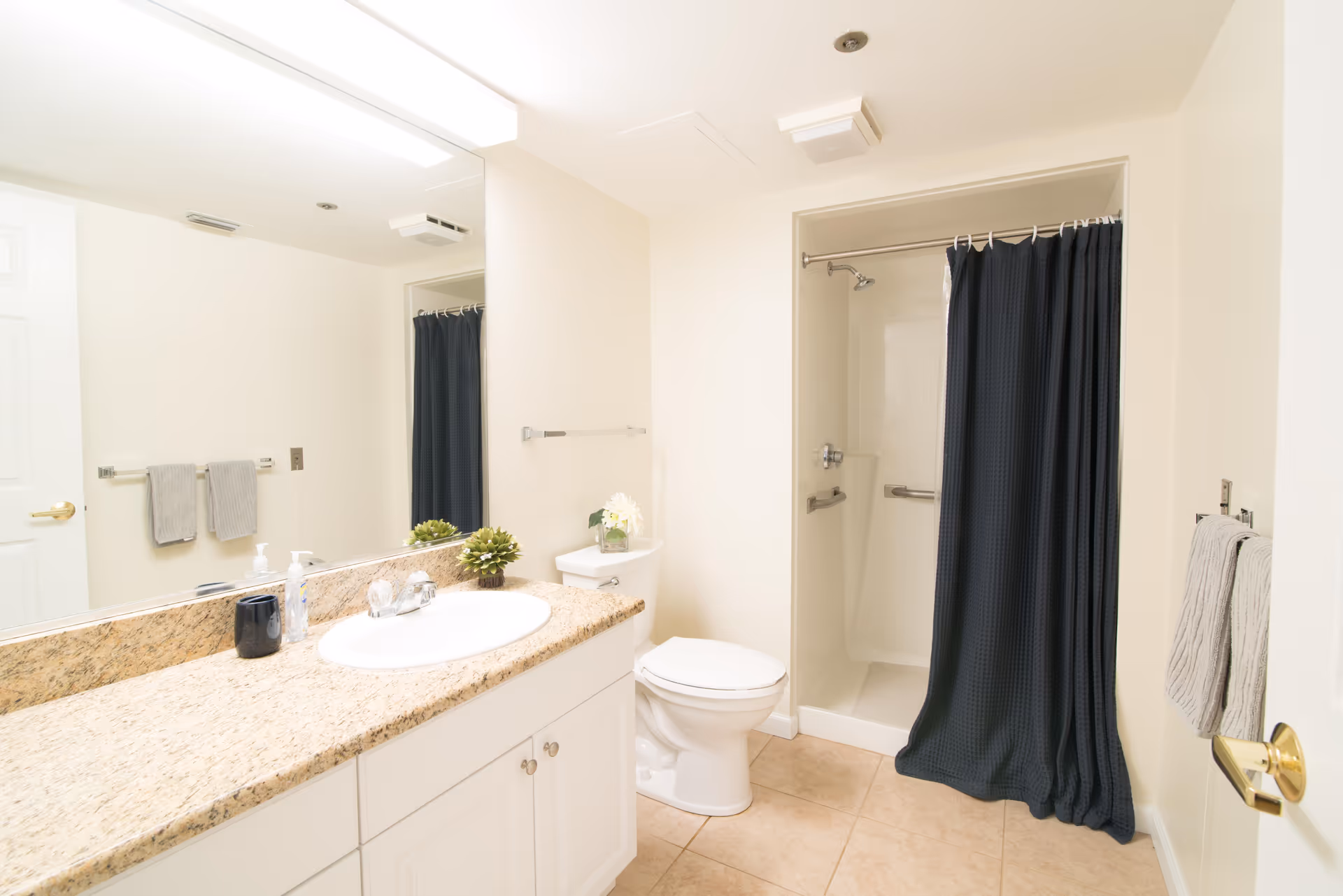 A clean and bright bathroom with a granite countertop sink, a toilet, and a shower with a dark blue curtain. The walls are light-colored, and there are two gray towels hanging on towel racks. A large mirror is above the sink, reflecting part of the room.