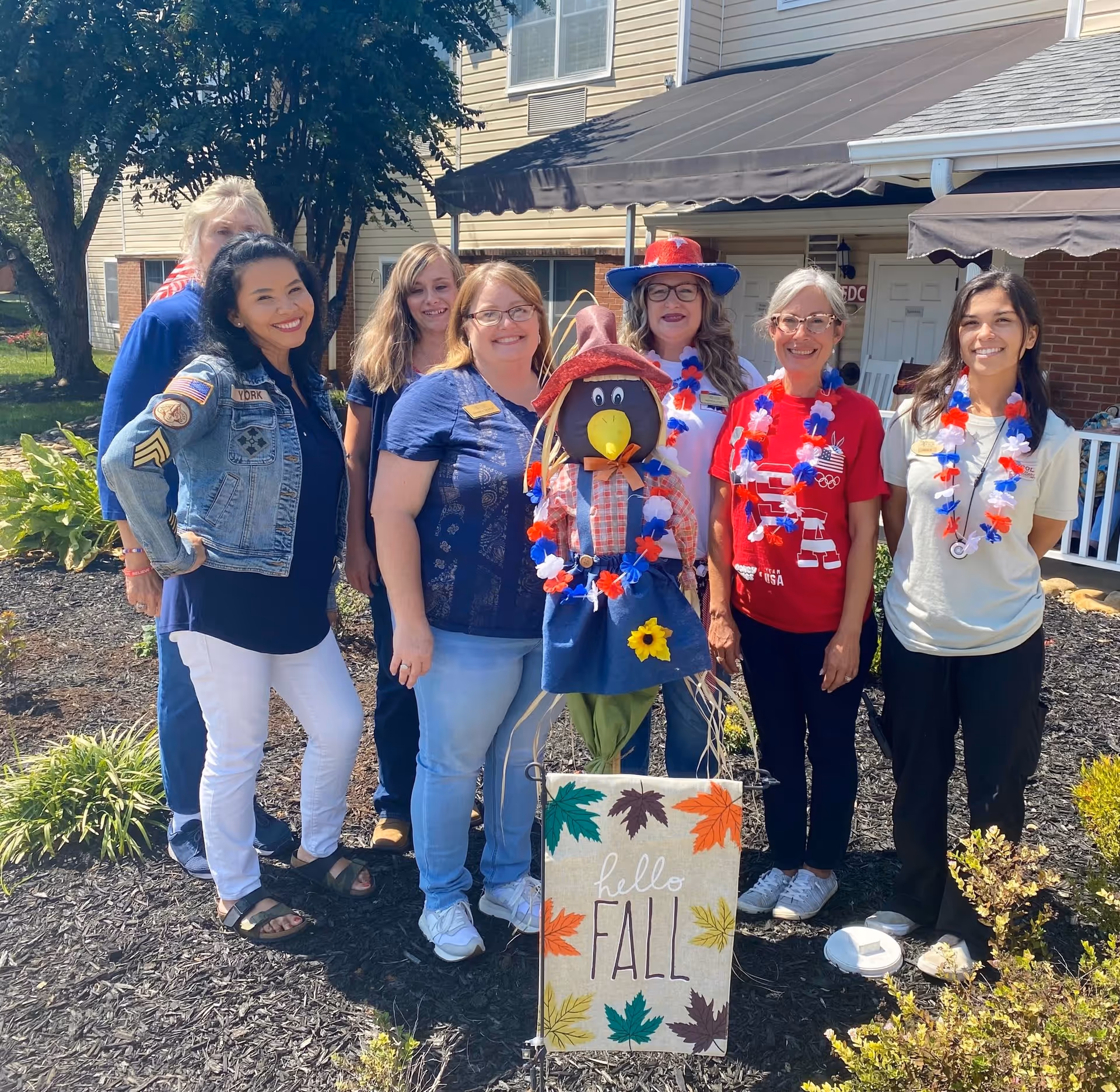 A group of six women standing outside in front of a building with a fall-themed scarecrow decoration and a sign that says 'hello FALL'. The women are smiling and some are wearing red, white, and blue leis. The background shows trees, plants, and part of the building with windows and awnings.