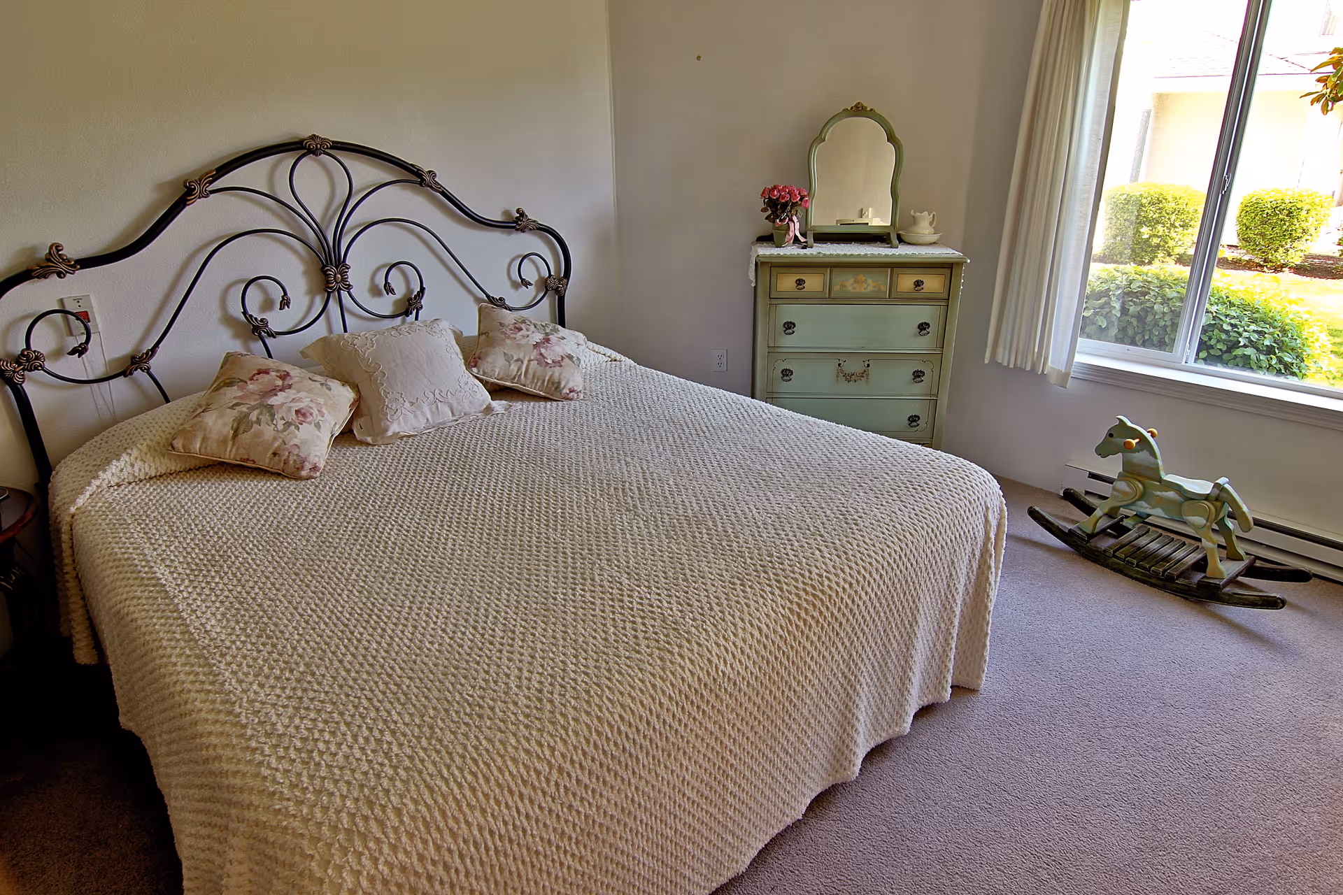 A bedroom with a large bed covered in a textured cream blanket and three decorative pillows. The bed has an ornate metal headboard. Next to the bed is a vintage green dresser with a mirror, a small vase with pink flowers, and a decorative item. A window with white curtains lets in natural light, showing green bushes outside. A wooden rocking horse is placed on the carpeted floor near the window.