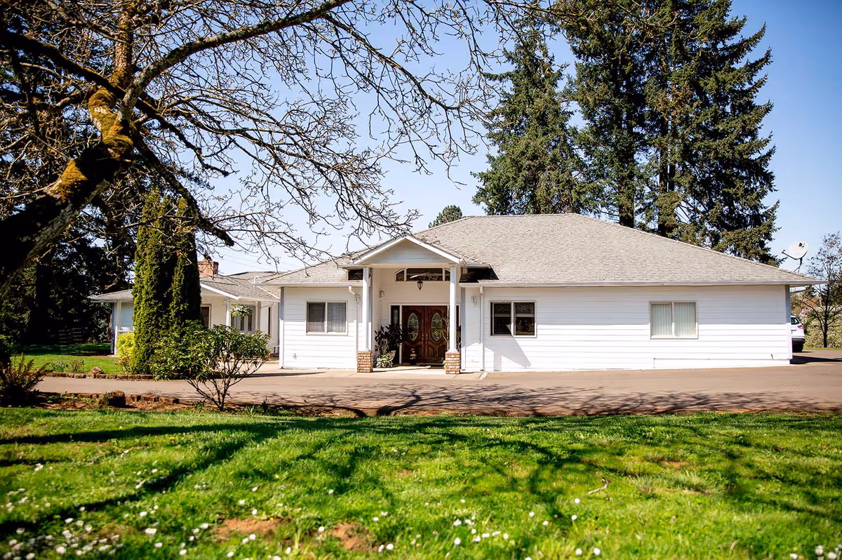 Front exterior of a single-story white house with a driveway, lawn, and surrounding trees.