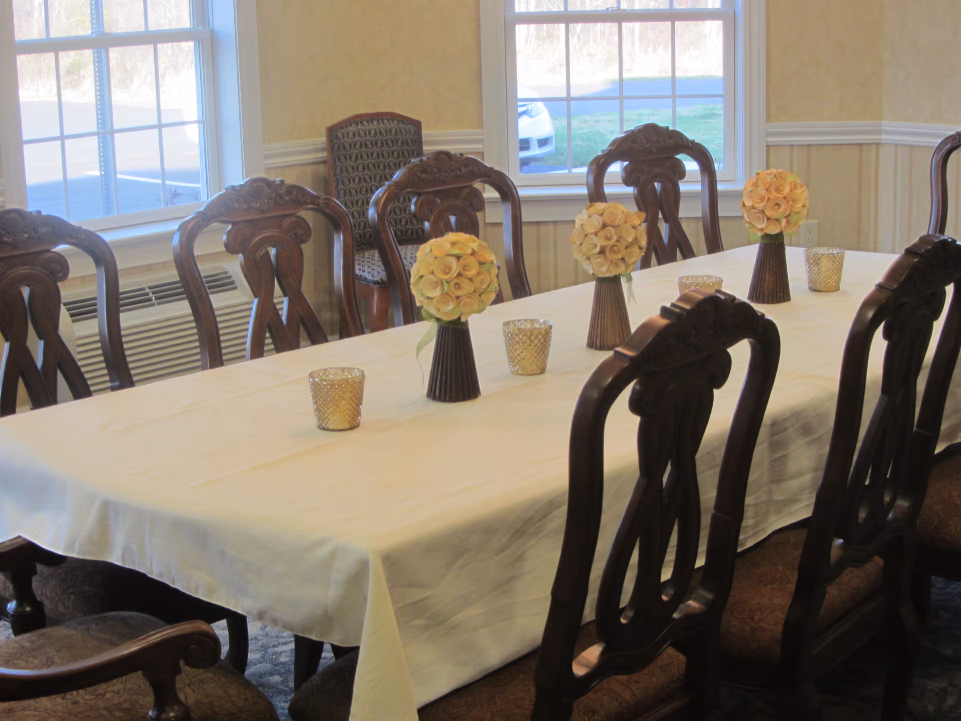 A dining room with a long rectangular table covered with a white tablecloth. The table is decorated with three vases of yellow roses and several small candle holders. The room has wooden chairs with ornate backs around the table and large windows letting in natural light.