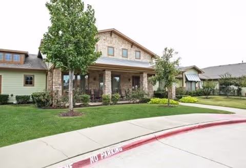 Front exterior view of a single-story assisted living facility with stone and wood siding, a covered porch with seating, green lawn, trees, and a curved driveway with a red-painted curb marked 'NO PARKING'.