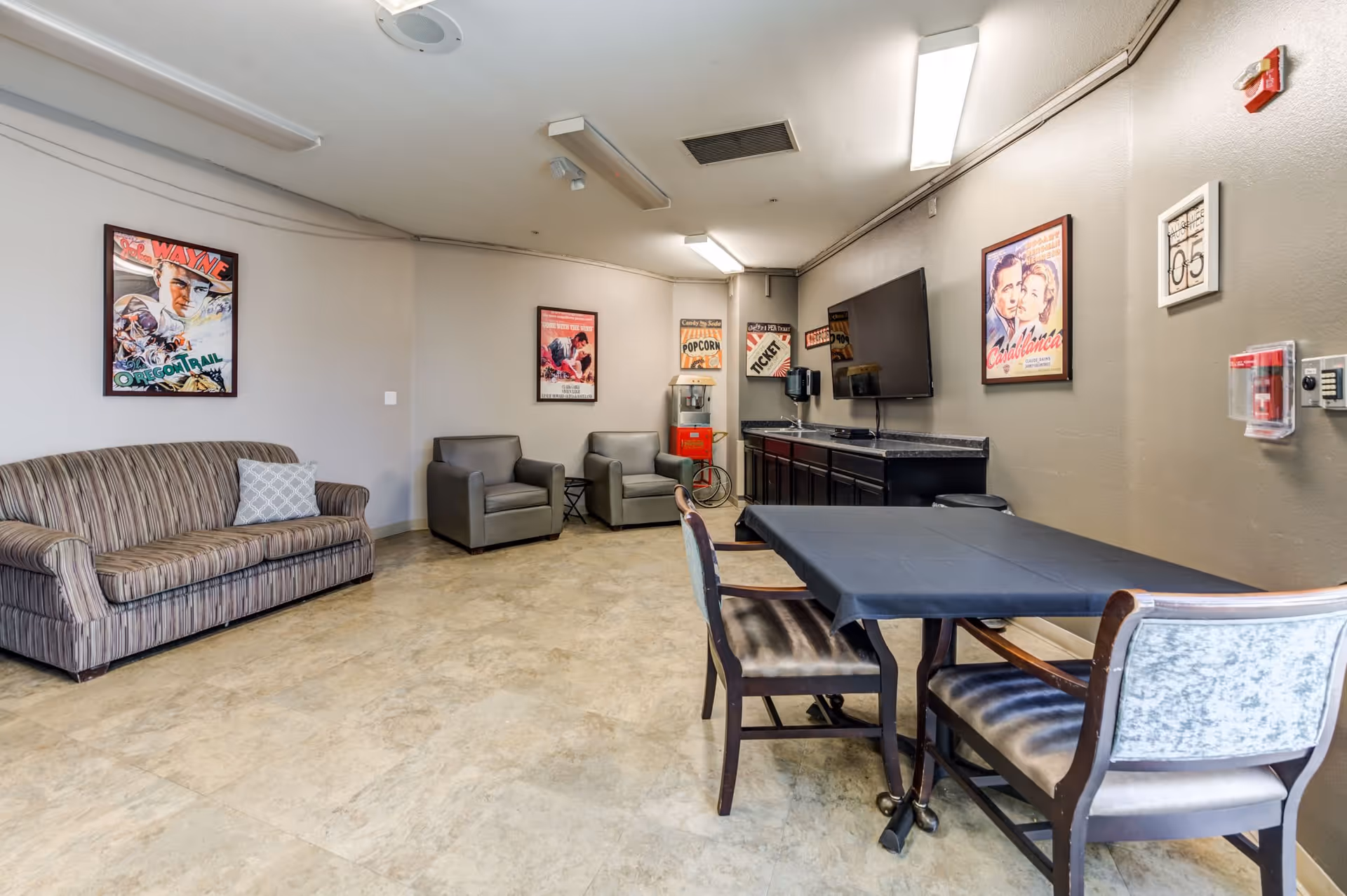 A cozy common area with a striped sofa, two gray armchairs, a table with four chairs covered with a black tablecloth, a wall-mounted TV, a popcorn machine, and vintage movie posters on the walls.