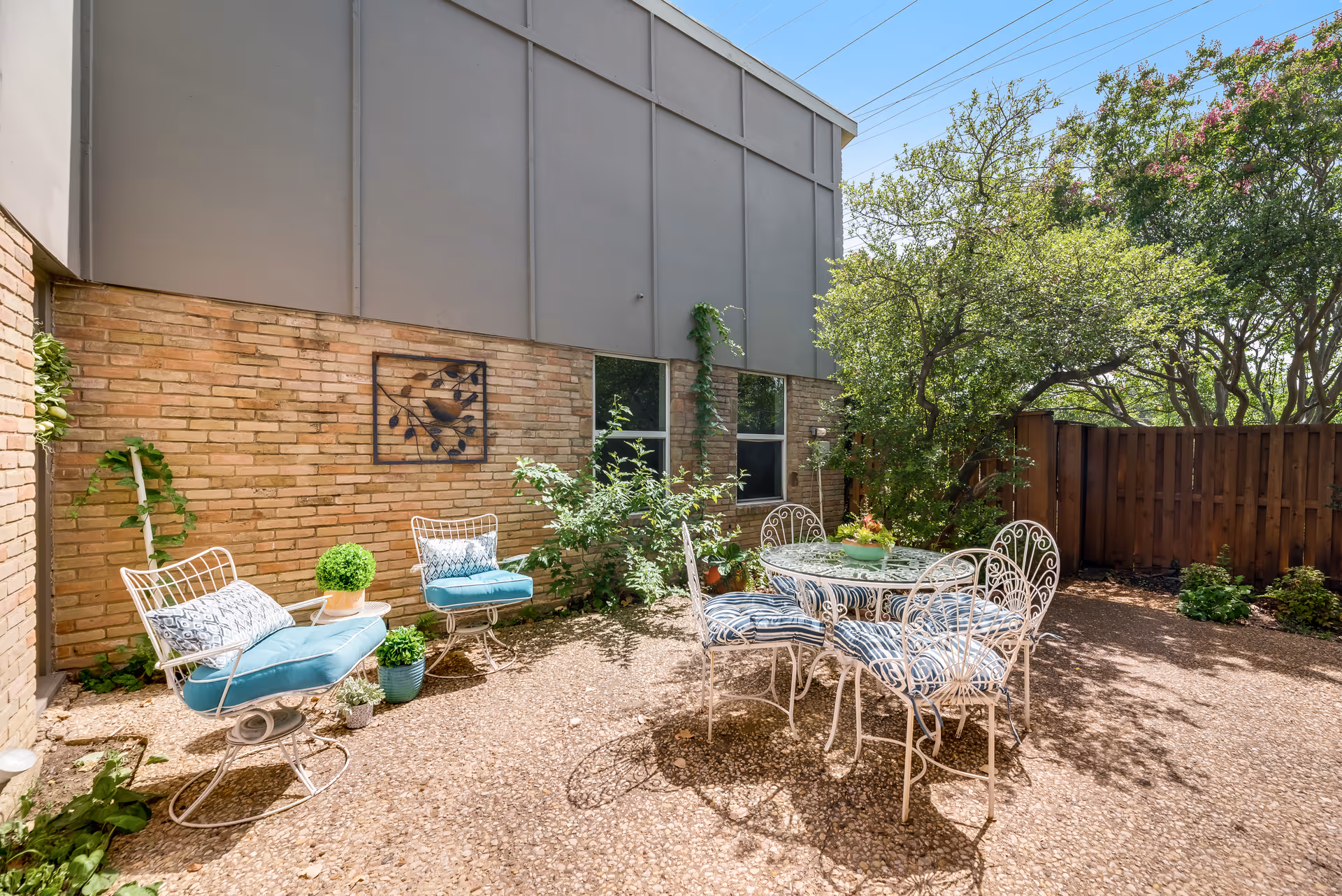A sunny outdoor patio area with a round glass-top table surrounded by four white metal chairs with blue and white striped cushions. Two white metal chairs with blue cushions and decorative pillows are placed near a brick wall with a metal wall art piece. The patio is surrounded by greenery including bushes and trees, and a wooden fence is visible in the background.