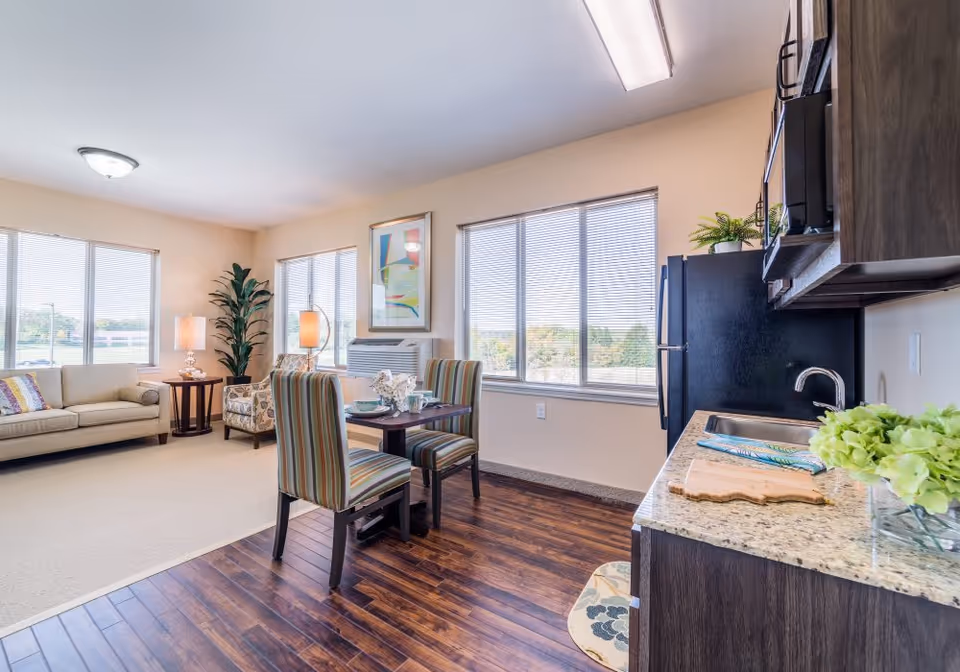 A bright and spacious senior living apartment featuring a small kitchen area with dark wood cabinets, granite countertops, and a sink. Adjacent to the kitchen is a dining area with a small table and two striped upholstered chairs. The living area includes a beige sofa, a patterned armchair, a side table with a lamp, and a tall potted plant. Large windows with blinds allow natural light to fill the room, and a colorful abstract painting hangs on the wall.