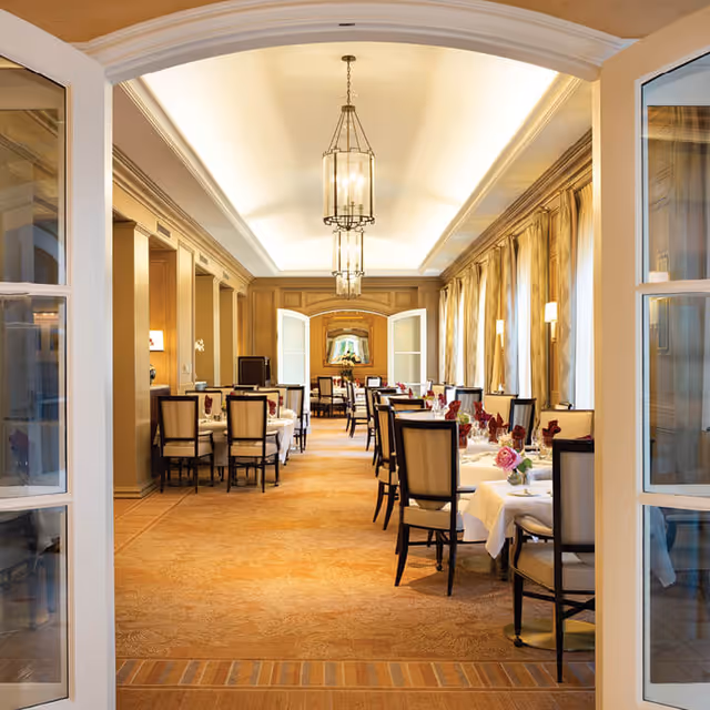 View through open double doors into an elegant dining room with tables set with white tablecloths, chairs, and floral centerpieces. The room features large windows with curtains on the right side, warm lighting from ceiling fixtures, and a carpeted floor.