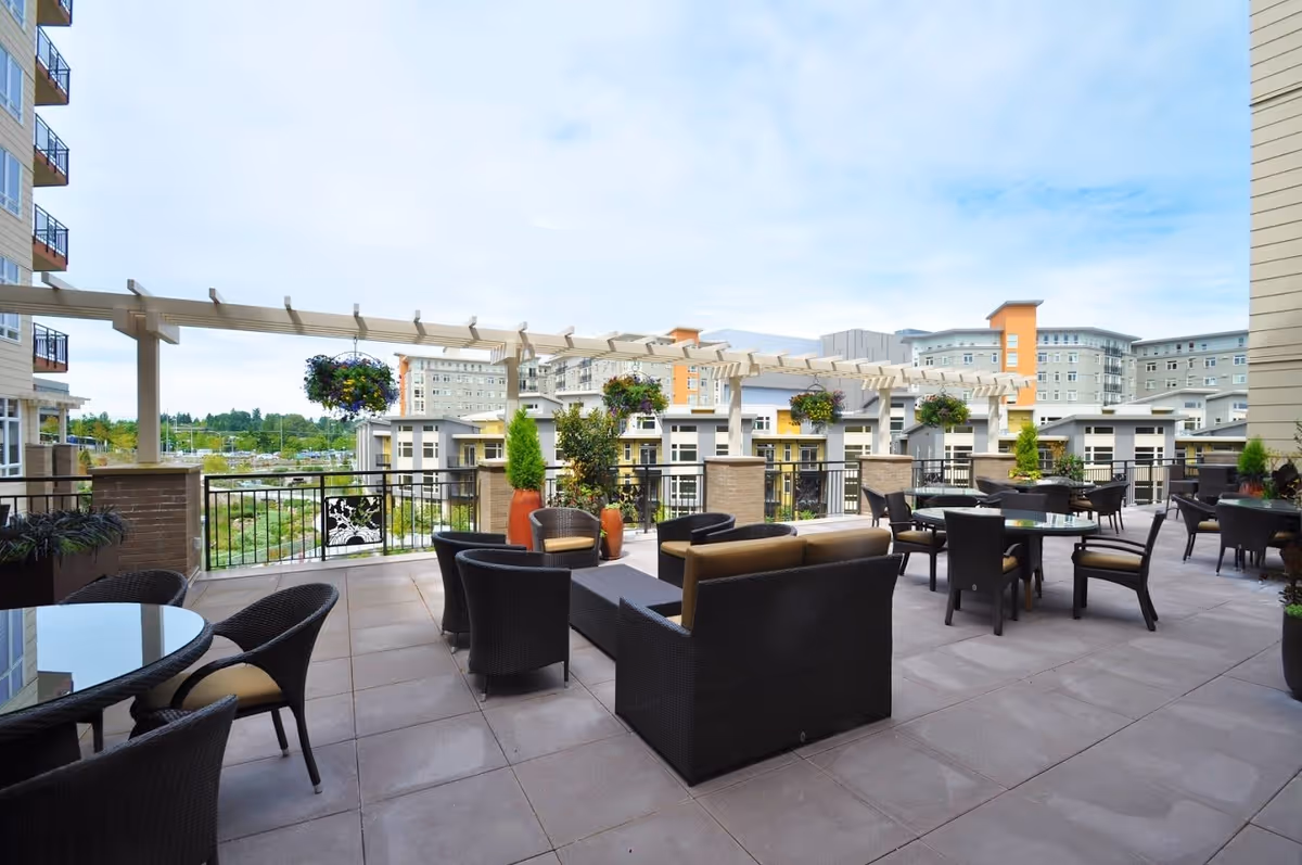 Outdoor patio area with multiple seating arrangements including wicker chairs and tables. There are hanging flower baskets and potted plants along the railing. In the background, there are multi-story residential buildings under a partly cloudy sky.