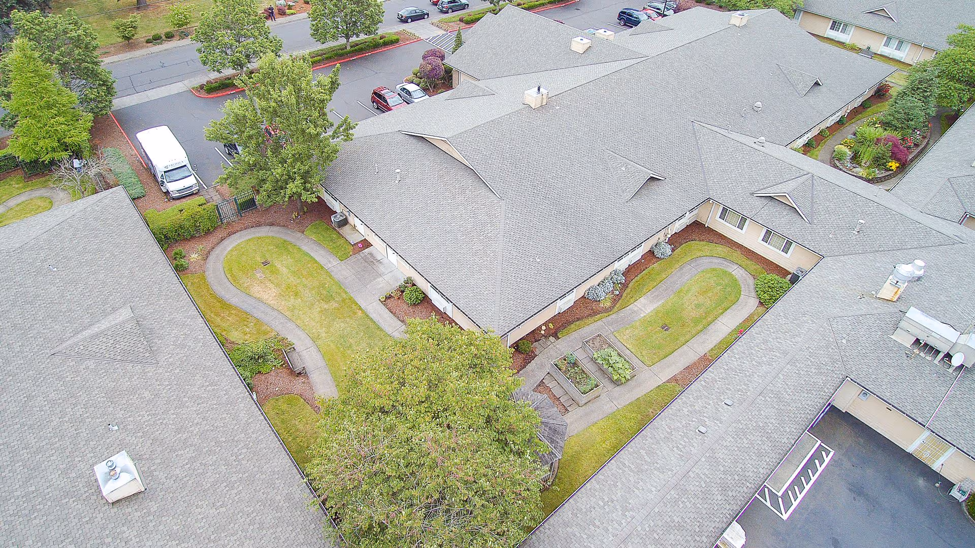 Aerial view of The Cottages Senior Living Memory Care facility showing multiple connected buildings with gray roofs surrounding a landscaped courtyard with walking paths, greenery, and garden beds. There are parked cars and a white van visible on the adjacent street and parking areas.