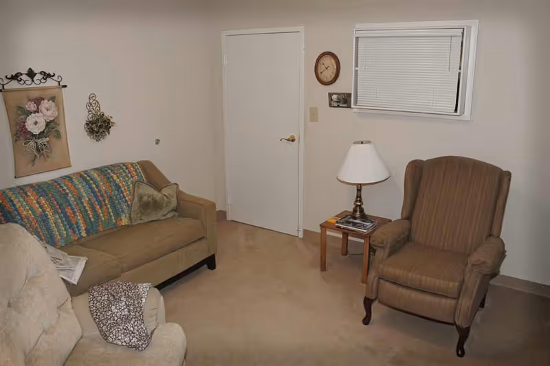 A small living room with beige carpet and light-colored walls. The room contains a brown sofa with a colorful crocheted blanket and a green pillow, a beige recliner with a patterned throw, and a brown striped armchair. There is a wooden side table with a lamp and some magazines, a wall clock, a small window with blinds, and floral wall decorations.