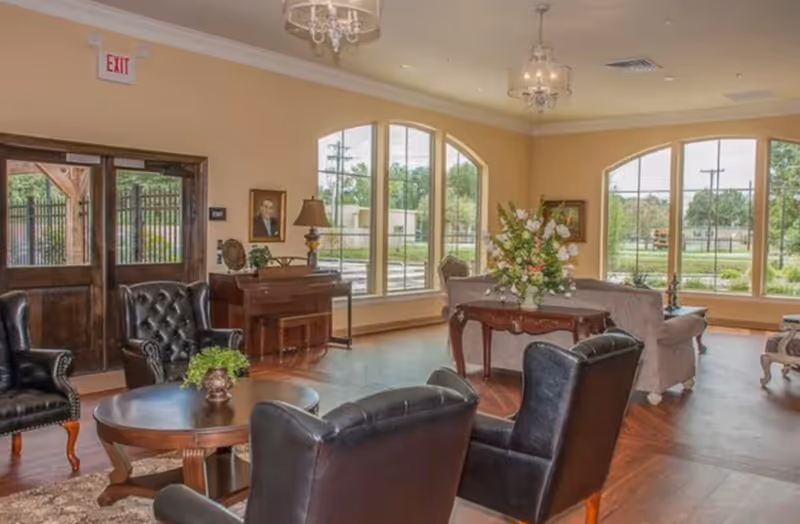 A spacious and well-lit living room area in Cedar Crest Memory Care featuring large arched windows, wooden floors, leather armchairs, a round wooden coffee table with a small plant, a piano against the wall, a table with a large floral arrangement, and elegant chandeliers hanging from the ceiling.
