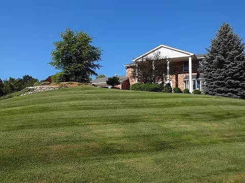 A large brick building with white columns partially visible behind a wide, well-maintained grassy hill with a few trees under a clear blue sky.