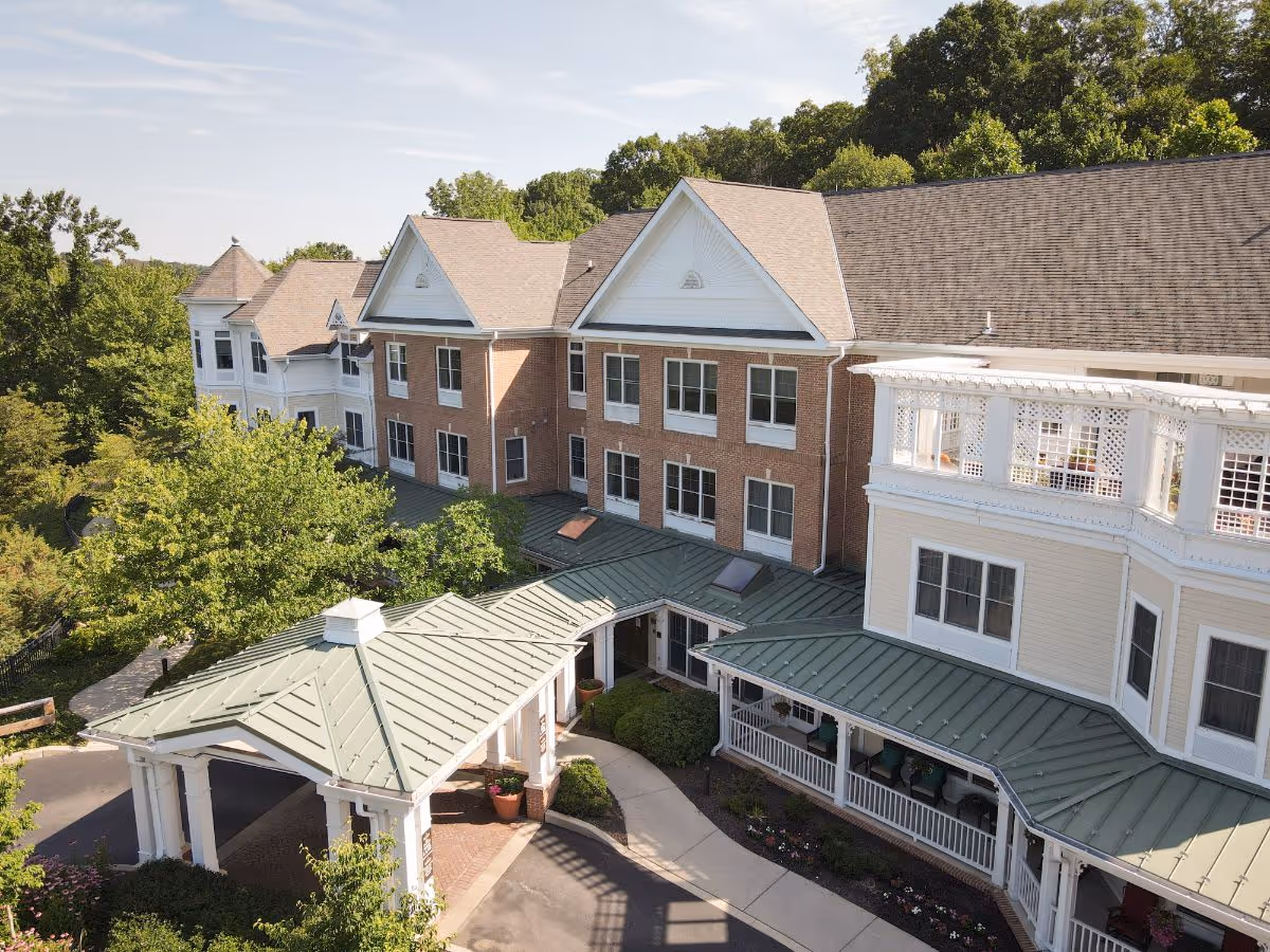 Exterior view of a senior living facility building with a covered entrance, multiple windows, and a porch area surrounded by trees and greenery under a clear sky.
