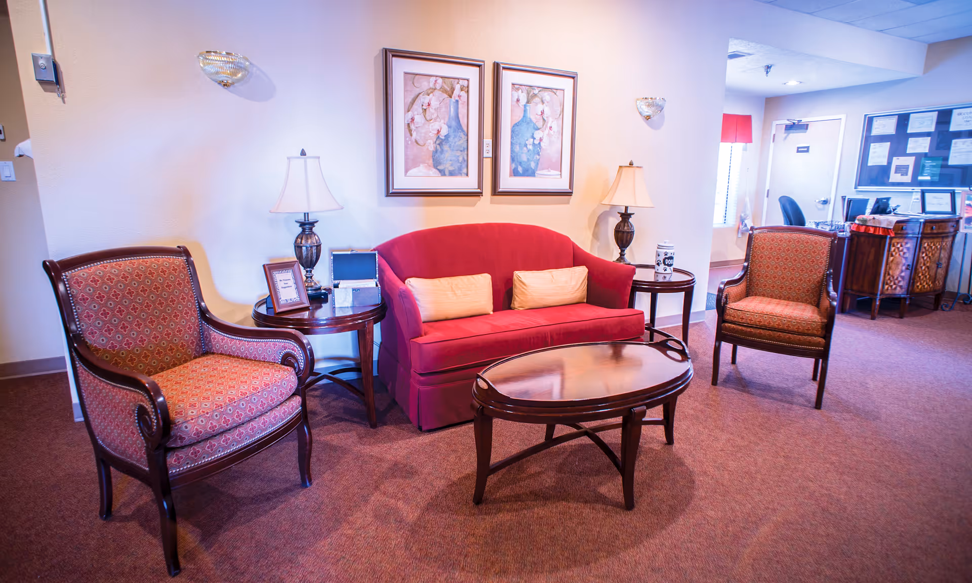 Cozy lobby seating area with a red loveseat, two patterned armchairs, wooden coffee table and side tables with lamps.