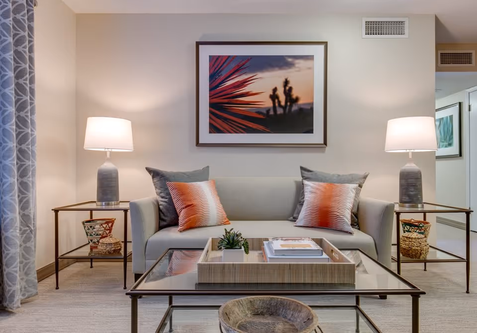 A cozy living room with a light gray sofa adorned with two gray and two orange patterned pillows. On either side of the sofa are glass-top side tables with gray lamps and decorative baskets underneath. A glass coffee table in front of the sofa holds a tray with books and a small plant. A framed picture of a desert scene with a sunset hangs on the wall above the sofa. Light-colored curtains with a geometric pattern are partially visible on the left side.