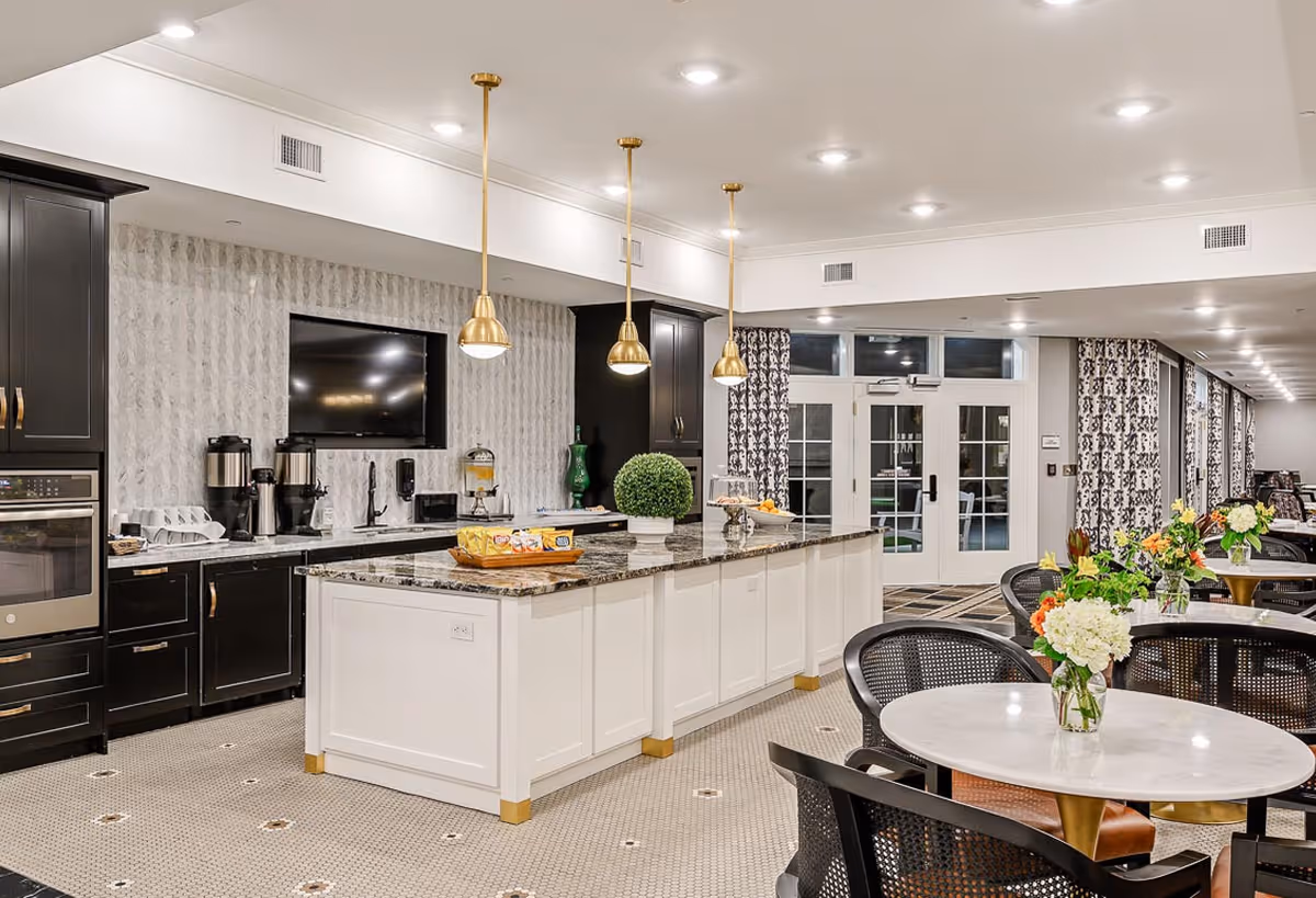 A modern dining area in a senior living facility featuring a large kitchen island with granite countertop and white cabinetry. The kitchen area has black cabinets, a built-in oven, coffee machines, and a mounted flat-screen TV. The dining space includes round tables with floral centerpieces and black chairs, with large windows and patterned curtains in the background.