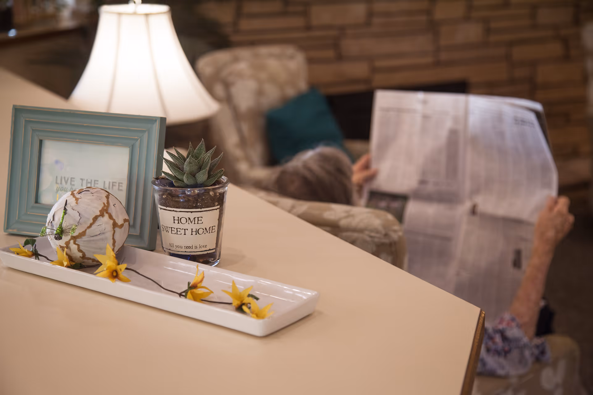 A close-up of a decorative arrangement on a table featuring a framed quote, a small potted succulent labeled 'Home Sweet Home', and a white tray with yellow flowers. In the background, an elderly person is sitting in a cushioned chair reading a newspaper in a cozy living room setting.
