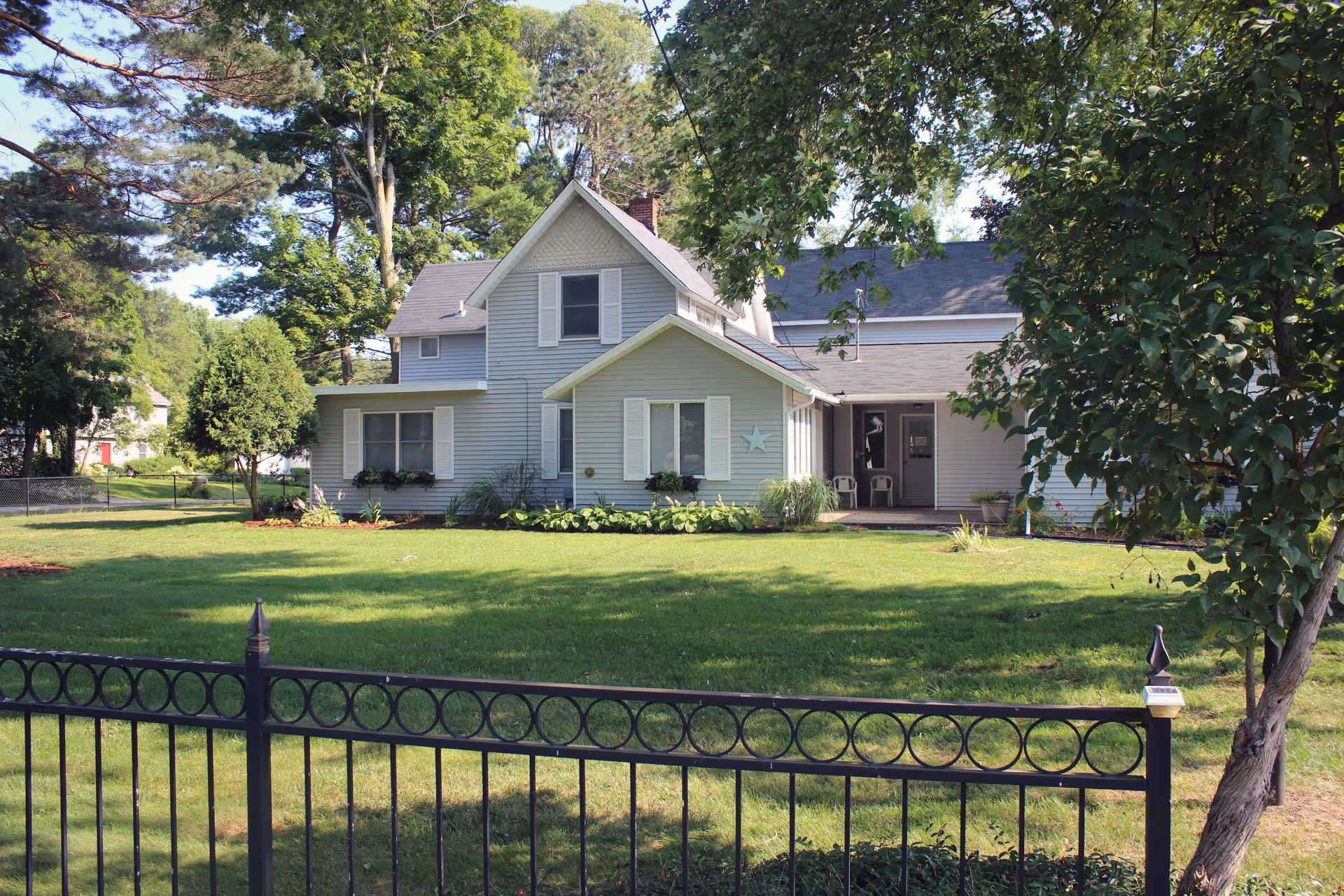 A light gray two-story house with white shutters and a star decoration on the front wall, surrounded by a well-maintained green lawn and trees, with a black metal fence in the foreground.