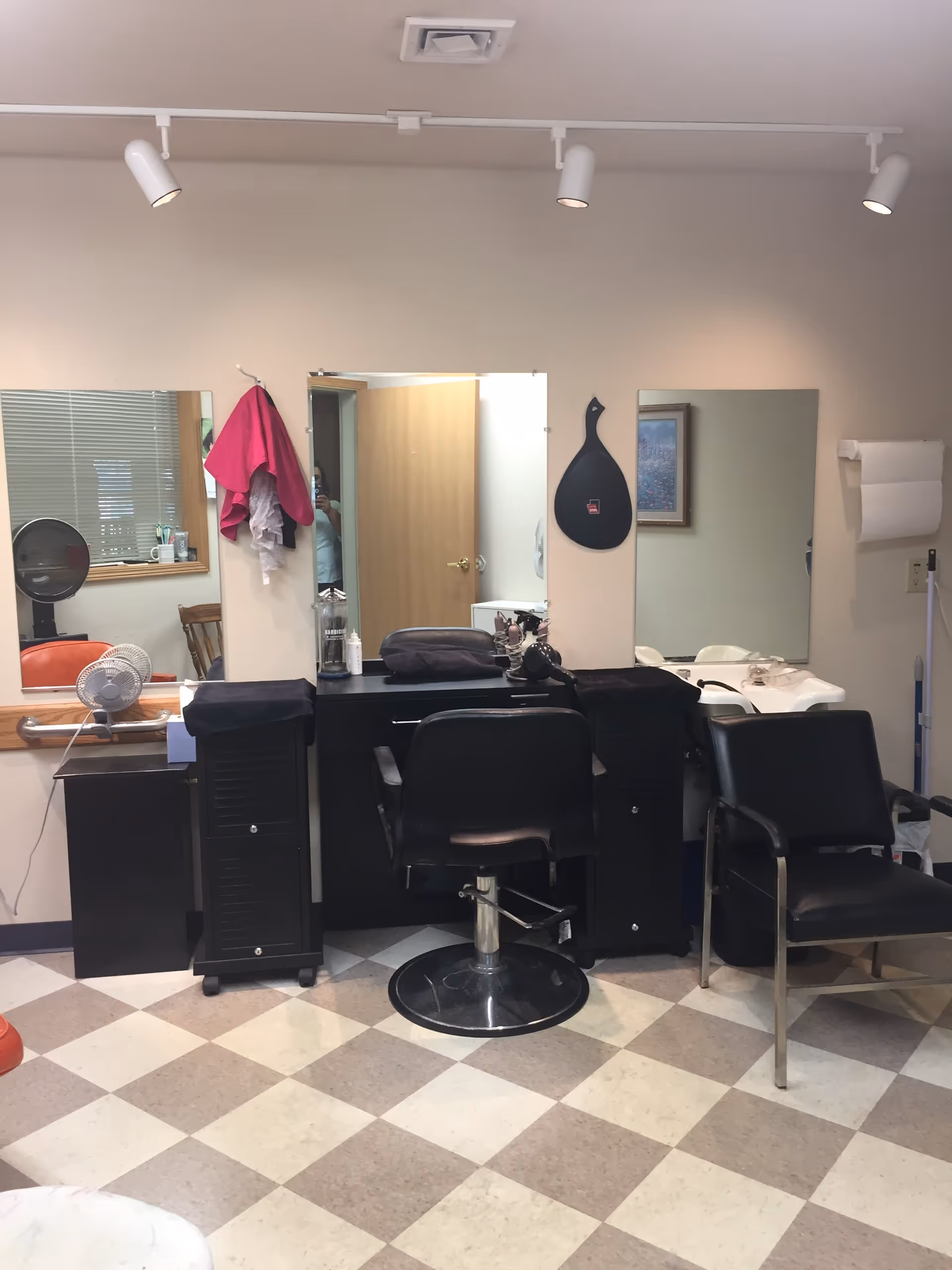 Interior view of a salon area with a black salon chair in front of a black counter with mirrors on the wall. There are two black cabinets on either side of the counter, a hair dryer, a handheld mirror hanging on the wall, and a black chair to the right. The floor has a checkered pattern with beige and light brown tiles.