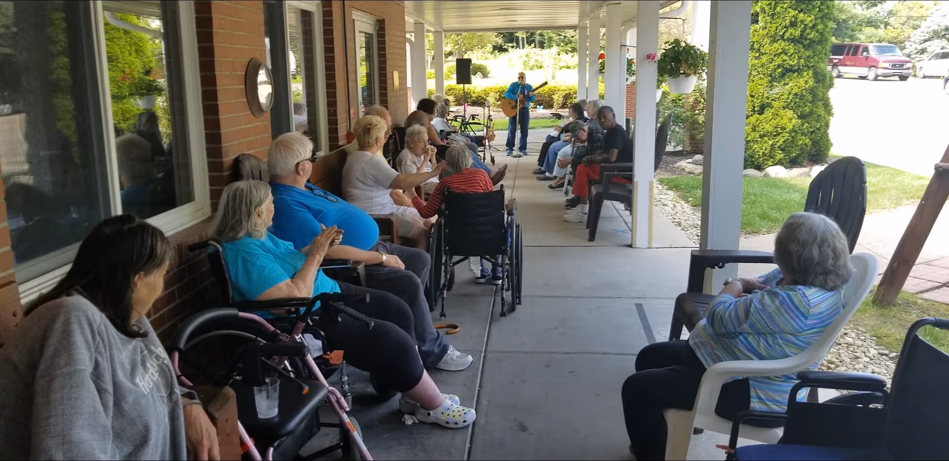 A group of elderly people sitting on chairs and wheelchairs along a covered outdoor walkway, attentively watching a person playing a guitar and singing at the far end. The setting is a sunny day with greenery and a parked vehicle visible in the background.