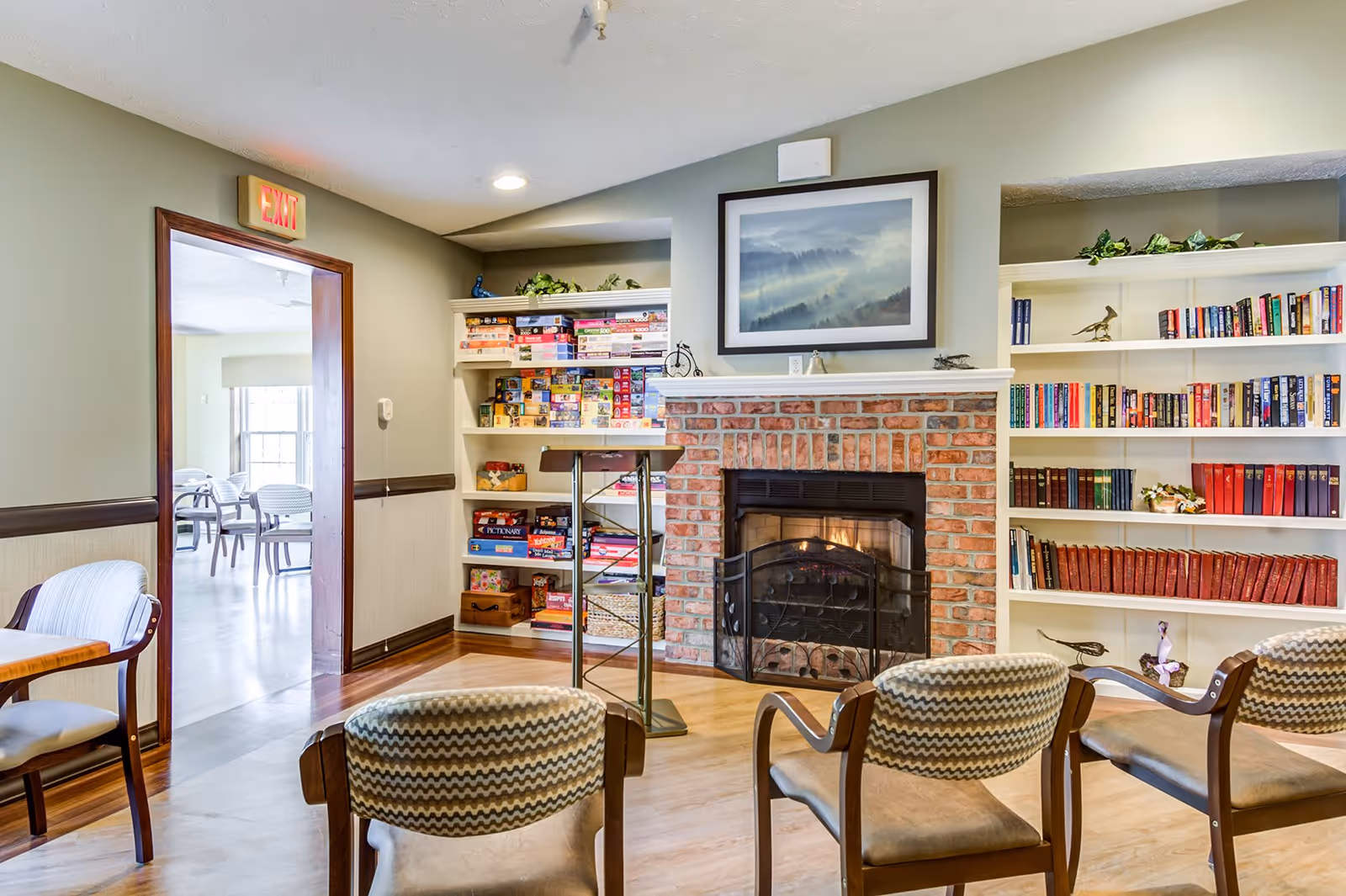 A cozy common area with a brick fireplace surrounded by white built-in bookshelves filled with books and board games. Three patterned chairs face the fireplace, and a wooden table with chairs is partially visible on the left. An exit sign is above a doorway leading to another room with more chairs and tables.