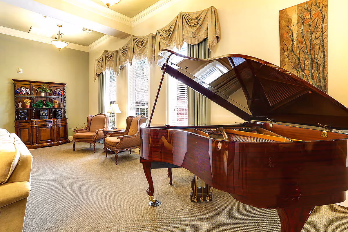 Bright lounge with a glossy grand piano, upholstered armchairs, a wooden cabinet, and tall windows with drapes.