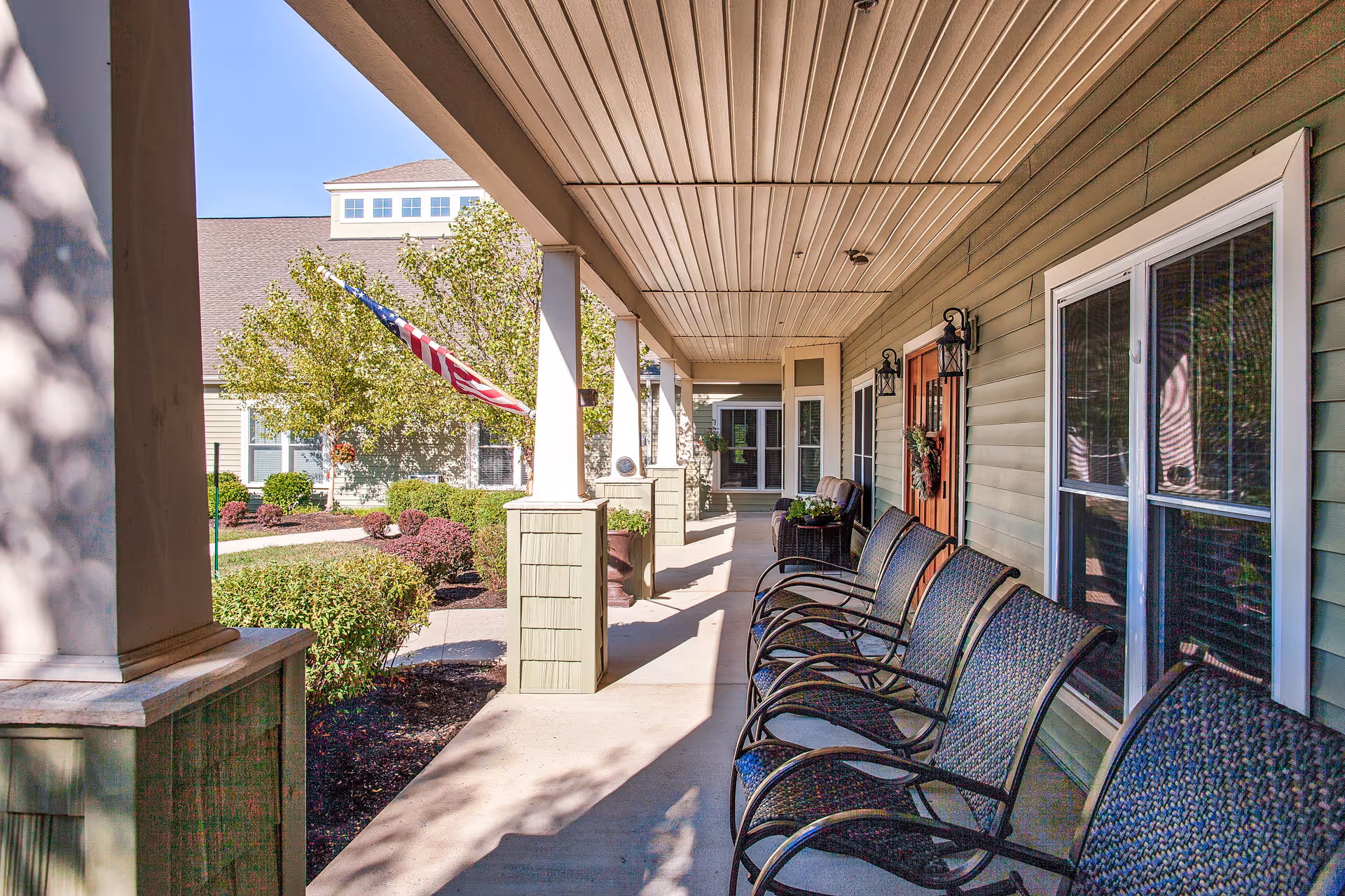Covered outdoor porch area with several black metal chairs lined up along the wall of a building. The porch has a beige ceiling and columns, with a view of a landscaped garden area featuring bushes, trees, and an American flag. The building has green siding and windows with white trim.