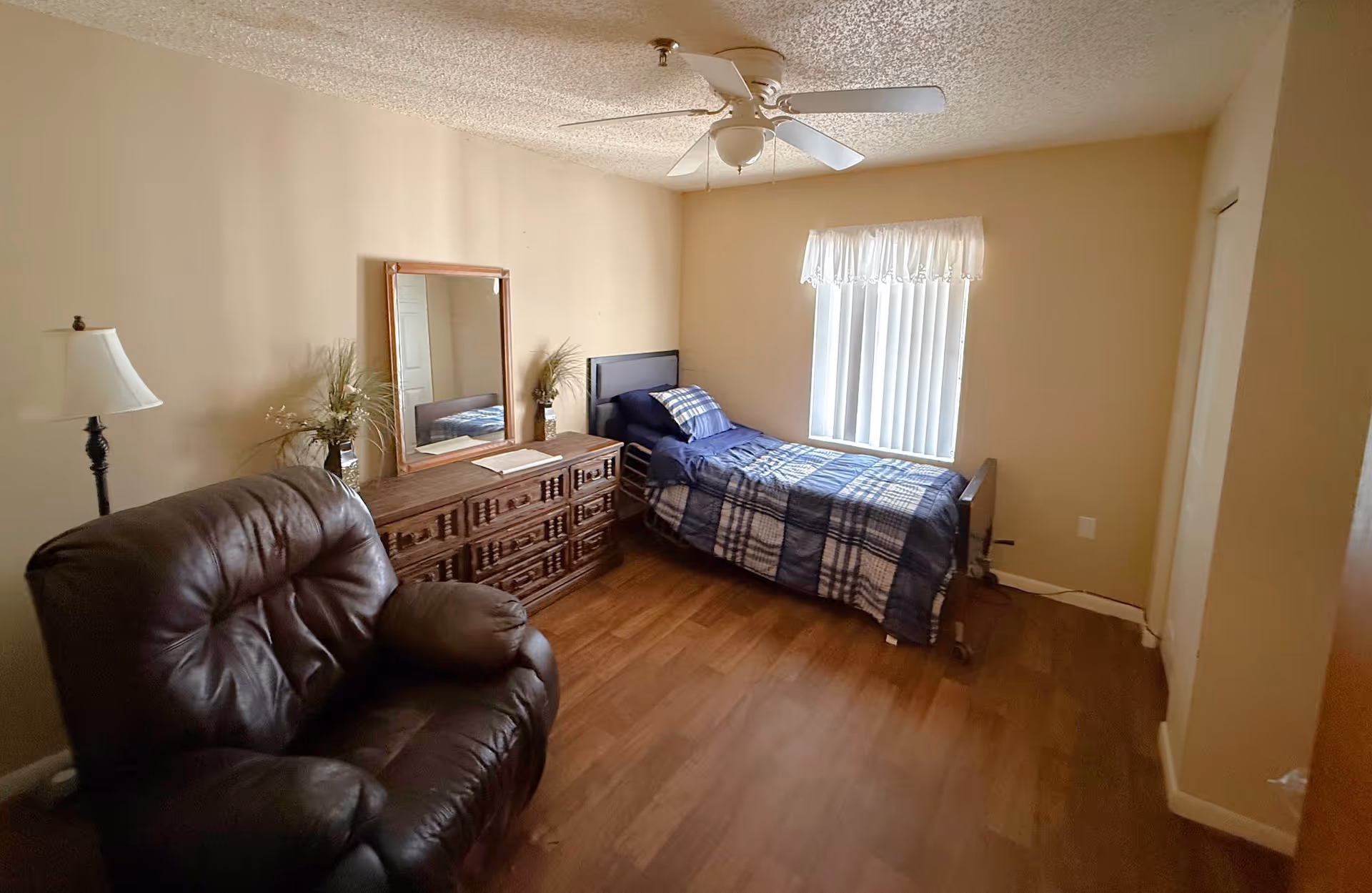 A small bedroom with a single bed covered in a blue and white plaid comforter, a wooden dresser with a mirror above it, a floor lamp, and a large brown leather armchair. The room has beige walls, a window with vertical blinds and a white valance, and a ceiling fan with a light fixture.