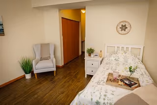 A cozy bedroom featuring a single bed with floral bedding, a white nightstand with a small plant and decorative items, a comfortable gray armchair with a cushion, a potted plant on the floor, and wooden flooring. The room has light-colored walls and a decorative wall hanging above the bed.