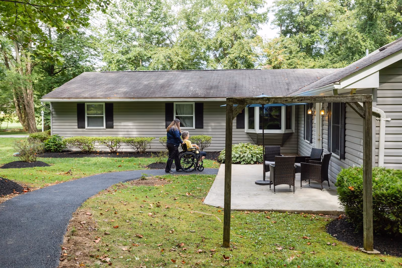 A caregiver pushes a person in a wheelchair along a paved path outside a single-story senior living building with a patio and outdoor seating.