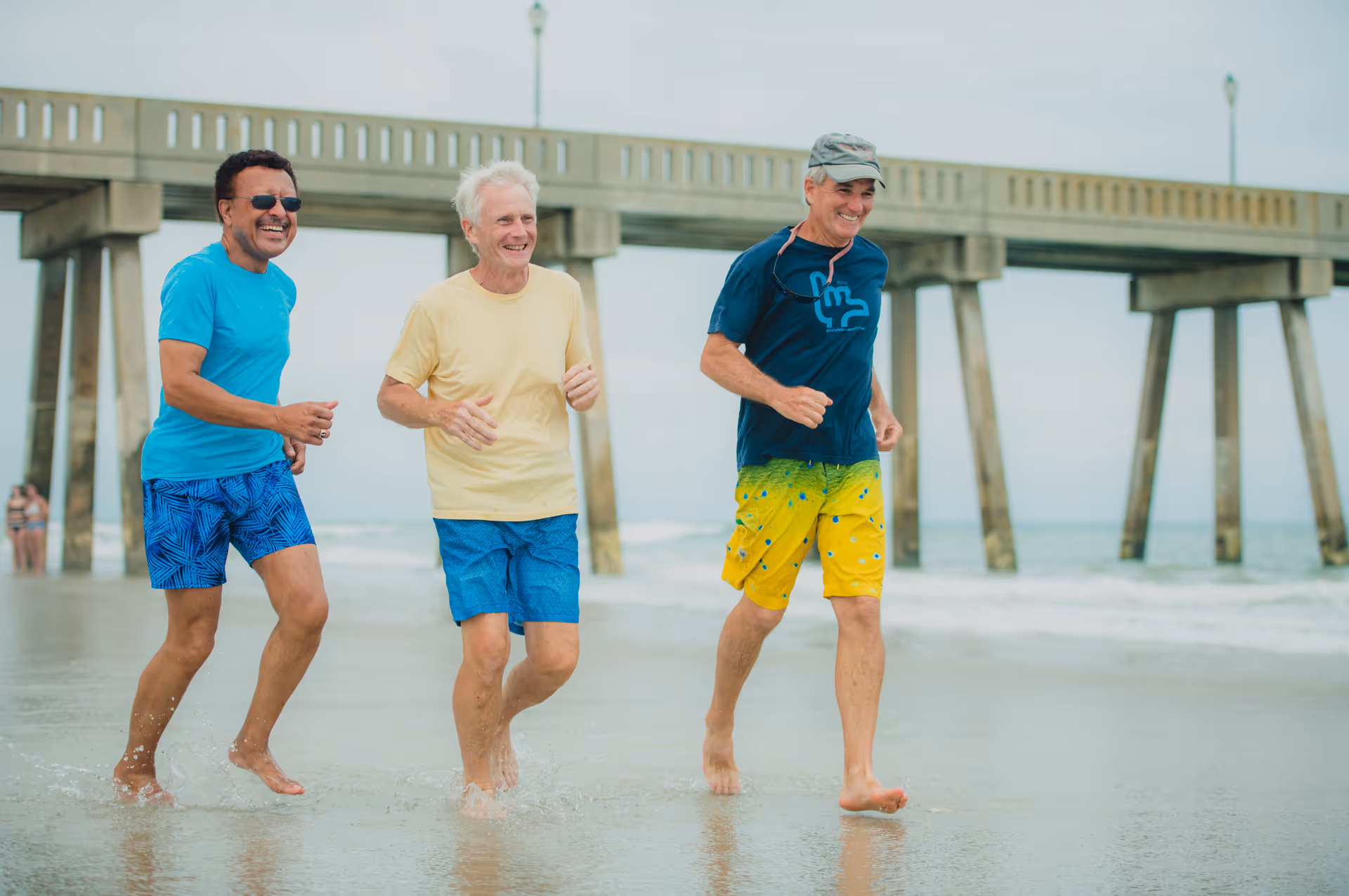 Three older men walking and smiling on a beach near a pier, with water and sand visible around their feet.