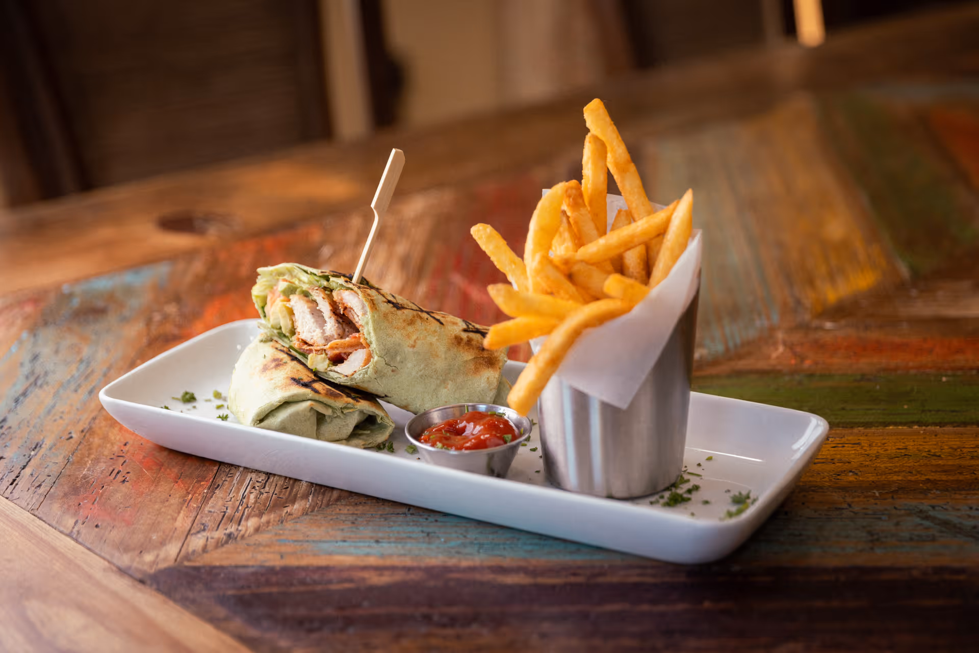 A plate with a wrap sandwich cut in half, a small metal cup filled with French fries, and a small container of ketchup on a rustic wooden table.