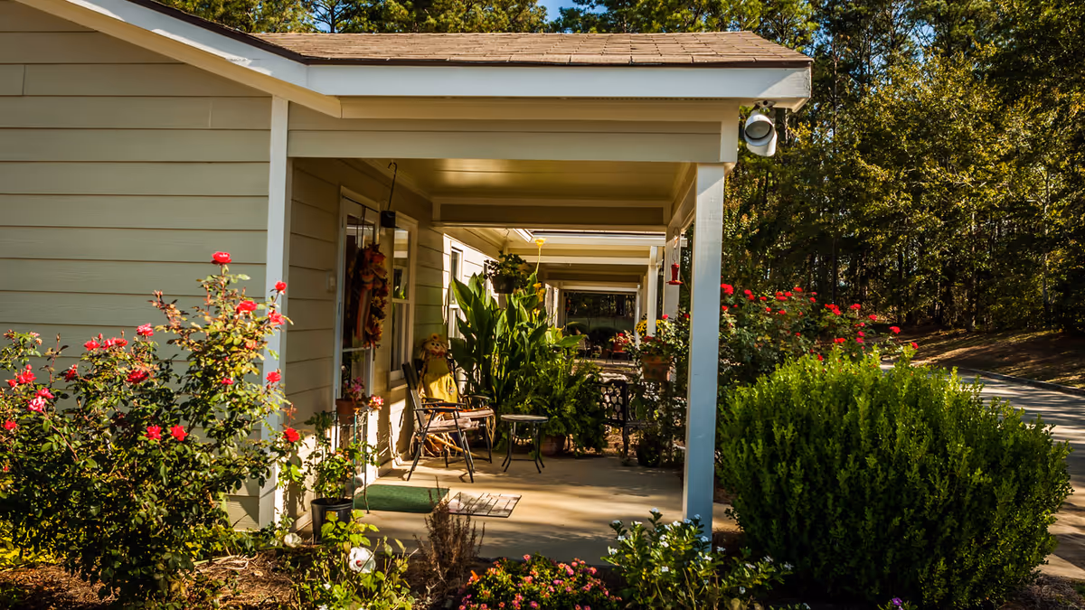 Covered front porch of an assisted living building with chairs, potted plants, and flower beds along the walkway.