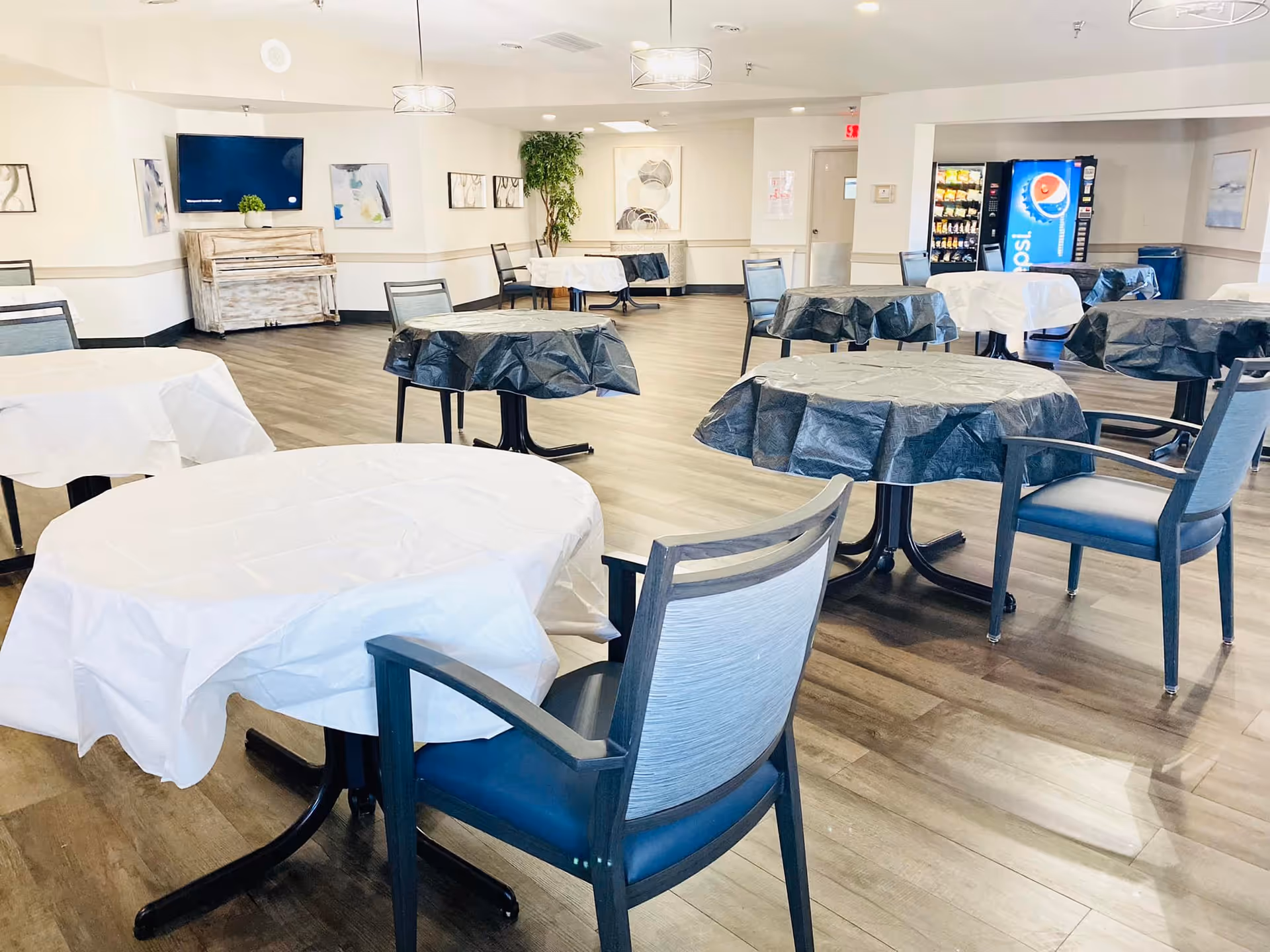 A spacious dining area with multiple round tables covered in black and white tablecloths, surrounded by chairs. The room has wood flooring, a wall-mounted TV, a piano, framed artwork on the walls, a vending machine, and a snack machine in the background.