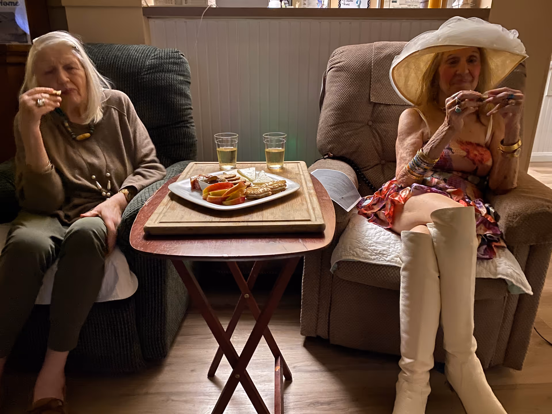Two elderly women sitting in recliner chairs in a cozy room. A small wooden table between them holds a plate with sliced apples, crackers, cheese, and two glasses of a light-colored beverage. One woman is wearing a brown sweater and green pants, while the other is dressed in a floral dress, white knee-high boots, and a large white hat. The background shows a white paneled wall with various items on a shelf above.