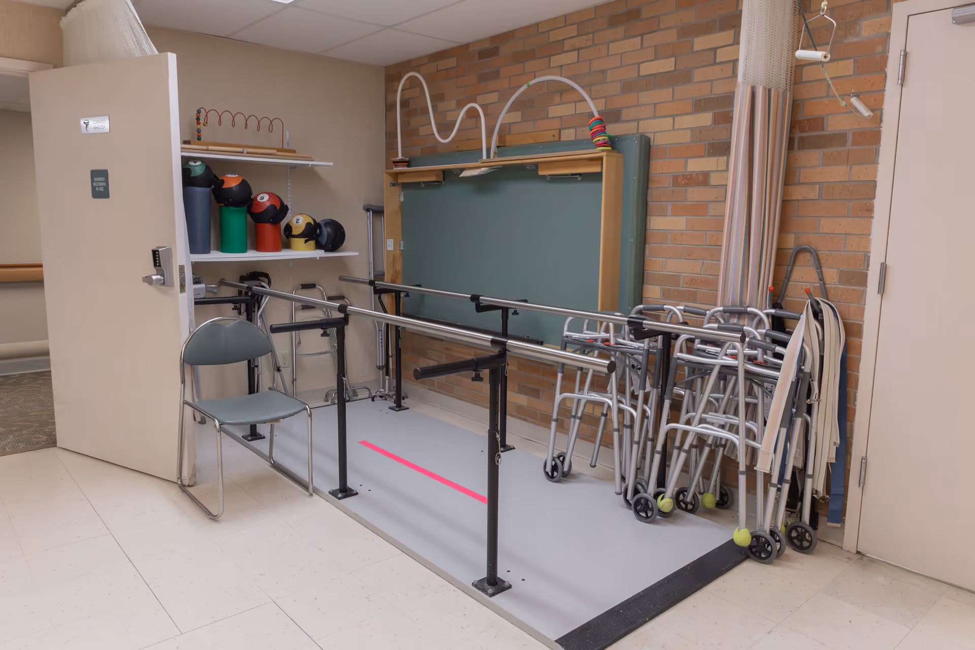 A physical therapy or rehabilitation room with parallel bars for walking exercises, several walkers lined up against a brick wall, a gray chair, and shelves holding colorful weighted balls and other therapy equipment.
