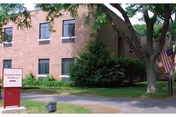 Exterior view of a brick building partially obscured by trees and bushes. There is a sign in front of the building that reads Elizabeth Seton Residence with an arrow pointing to the left. An American flag is visible near the right side of the image.