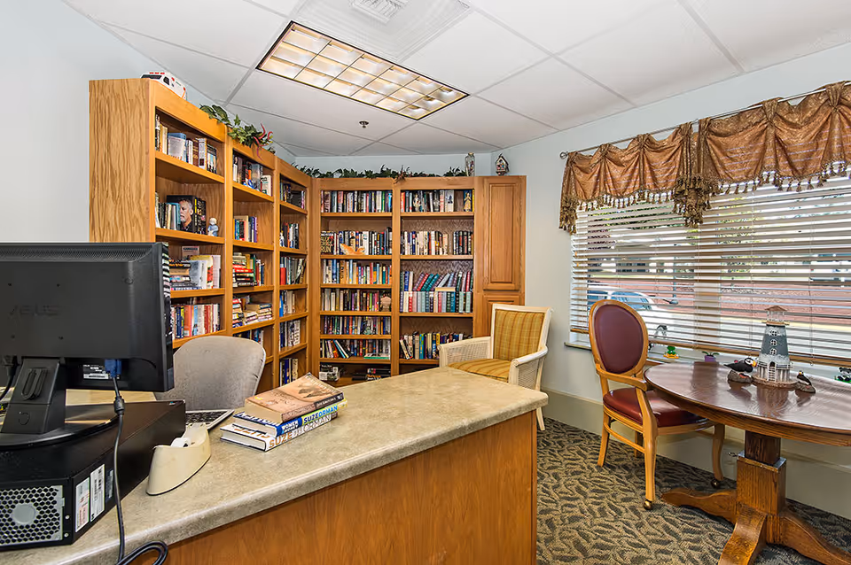Interior view of a cozy reading room in a retirement community featuring wooden bookshelves filled with books, a desk with a computer and books on it, two chairs, and a round table near a window with blinds and decorative curtains.
