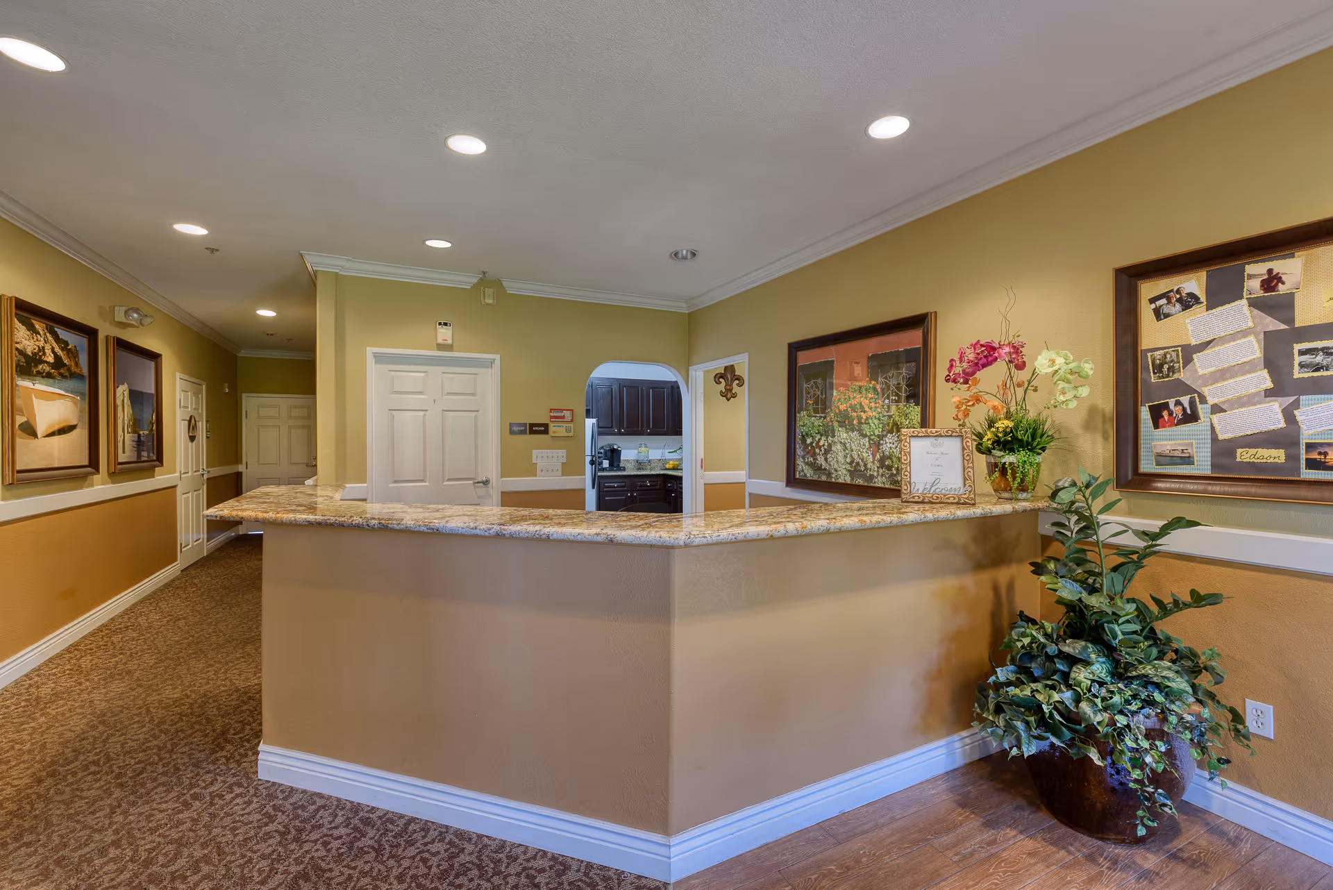 Reception area with a granite countertop desk, beige walls, and carpeted floor. The wall behind the desk has a door and an open doorway leading to a kitchen area with dark cabinets. The walls are decorated with framed pictures and a bulletin board. A large potted plant and a vase with flowers are placed near the desk.