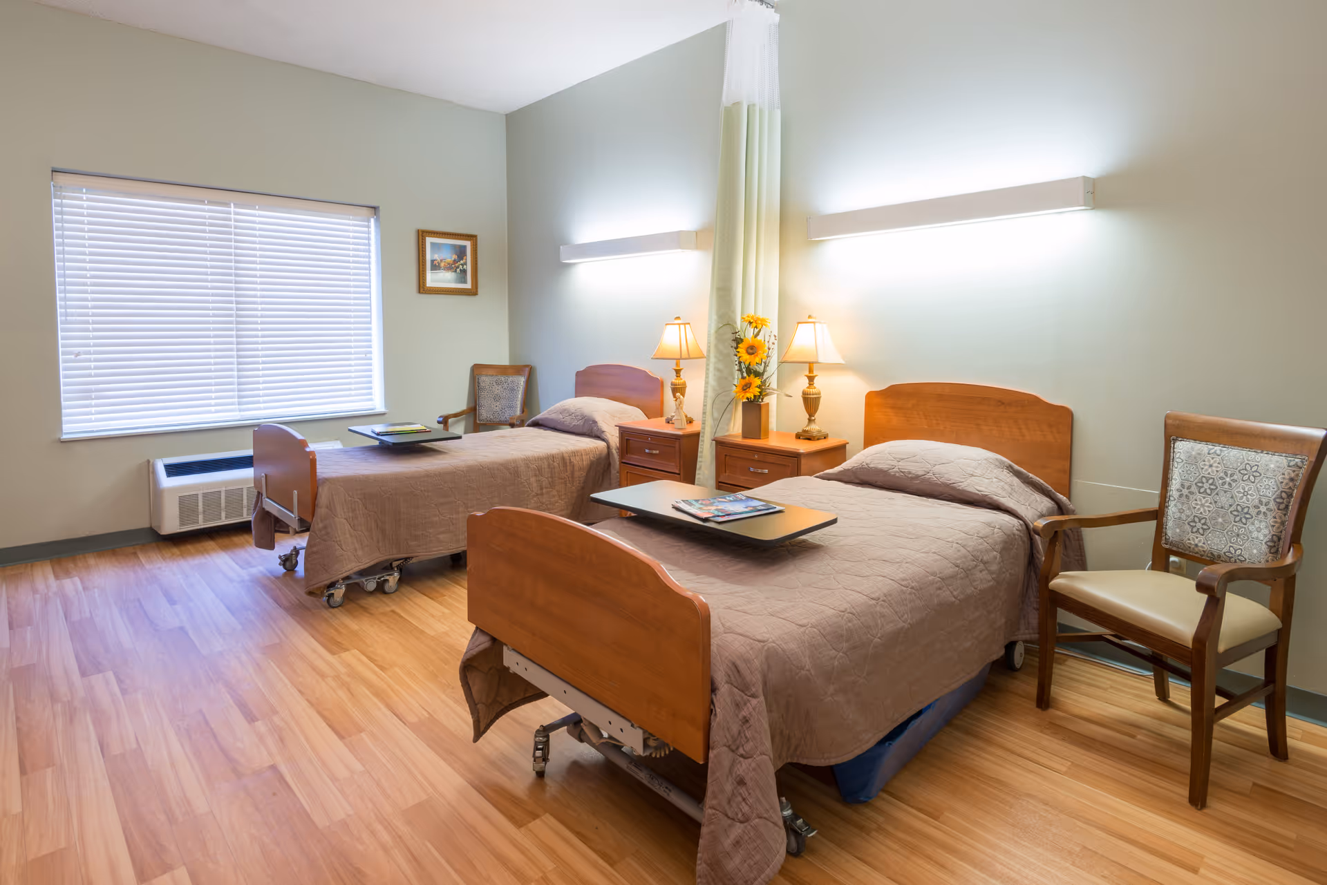 A bright and clean room with two single hospital-style beds, each with a brown quilted bedspread. Between the beds are two wooden nightstands with lamps and a vase of sunflowers. There are two wooden chairs with patterned cushions, a large window with closed blinds, and a light green wall with mounted lights above each bed. The floor is wood laminate.