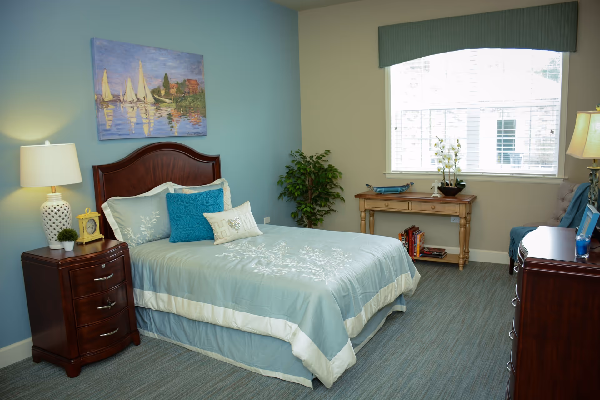 A neatly made bed with light blue and white bedding in a bedroom. The bed has a dark wooden headboard and is flanked by a wooden nightstand with a lamp, a small plant, and a clock. There is a painting of sailboats on the wall above the bed. A window with blinds lets in natural light, and a wooden table with books and a plant is placed against the wall. The room has a blue carpet and a comfortable, inviting atmosphere.